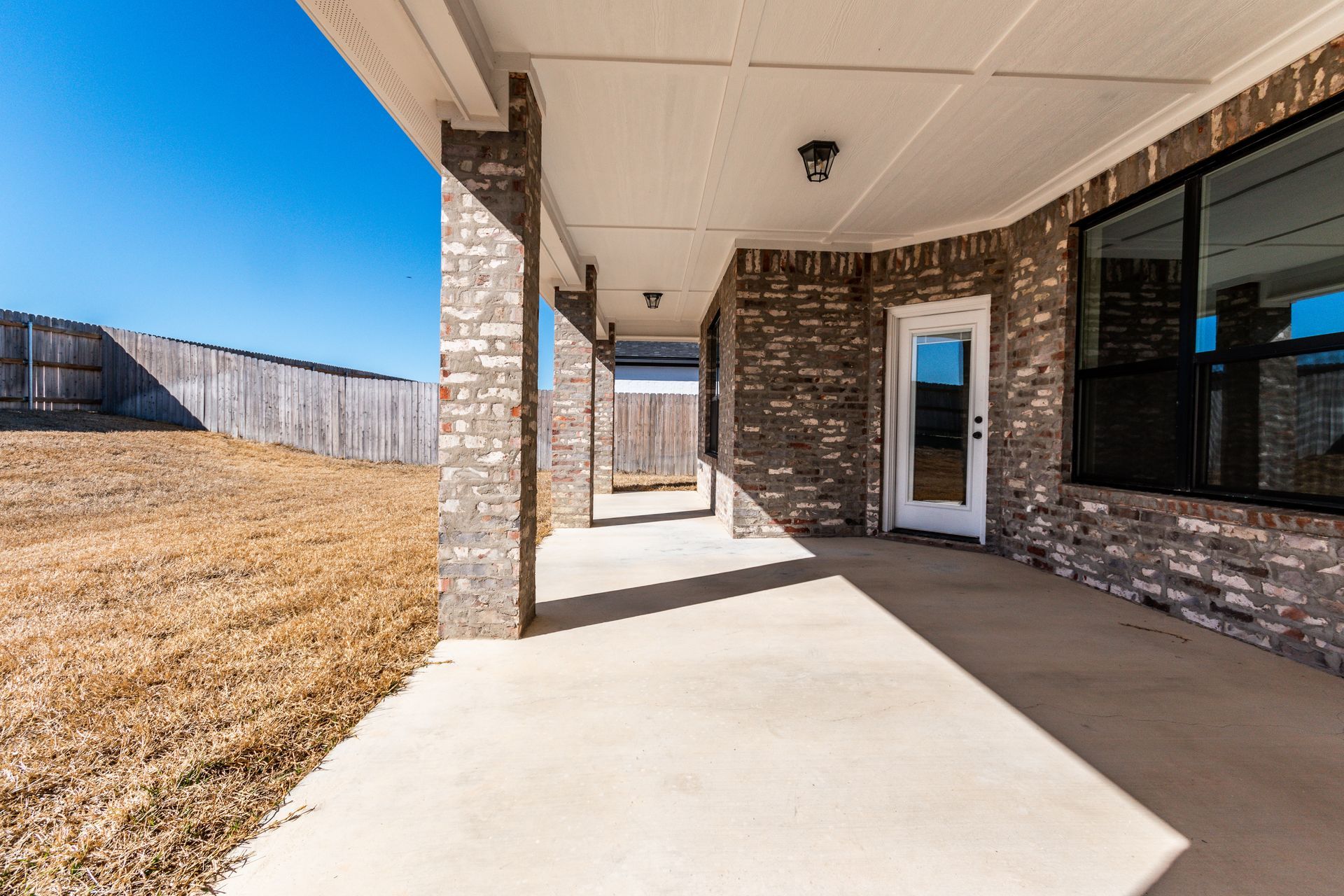 Covered patio with brick columns, concrete floor, and a door, next to a backyard.