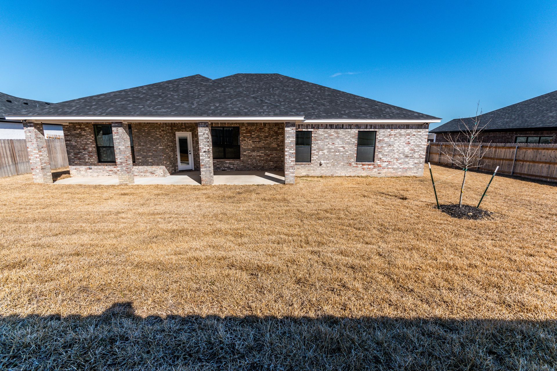 Backyard of a brick house with a brown, dry lawn under a clear blue sky.