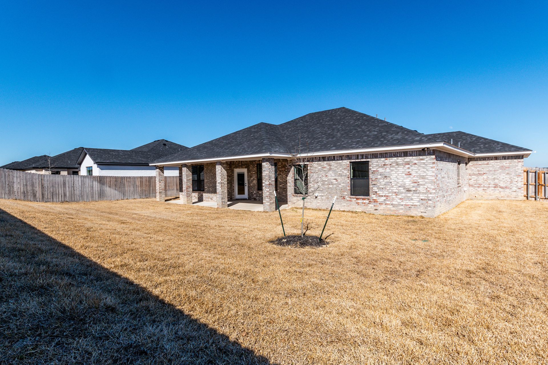 Brick house with black roof, brown yard, wooden fence, and blue sky.