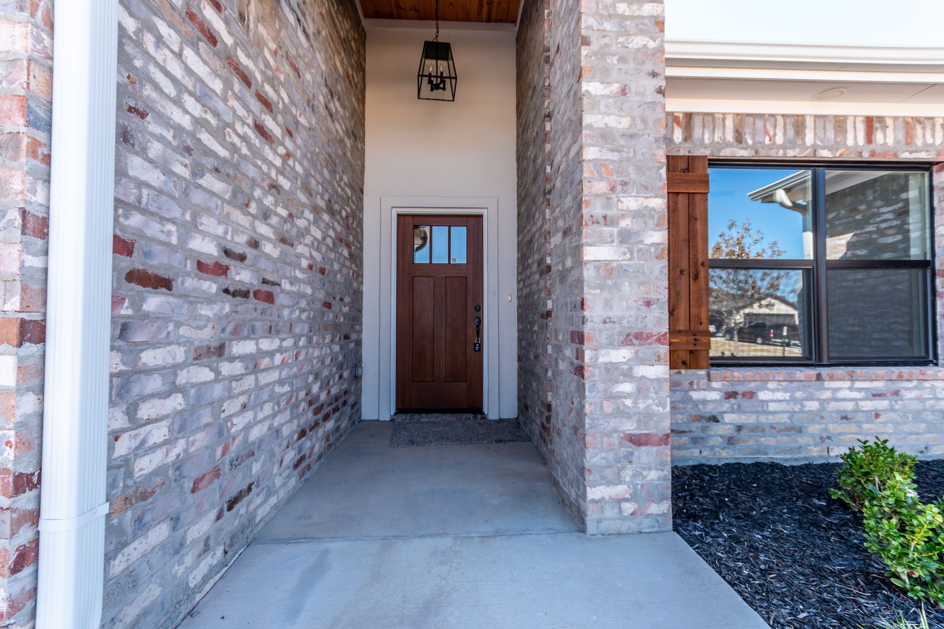 Exterior of a brick home with brown door under a covered entryway. Window with shutters to the right.