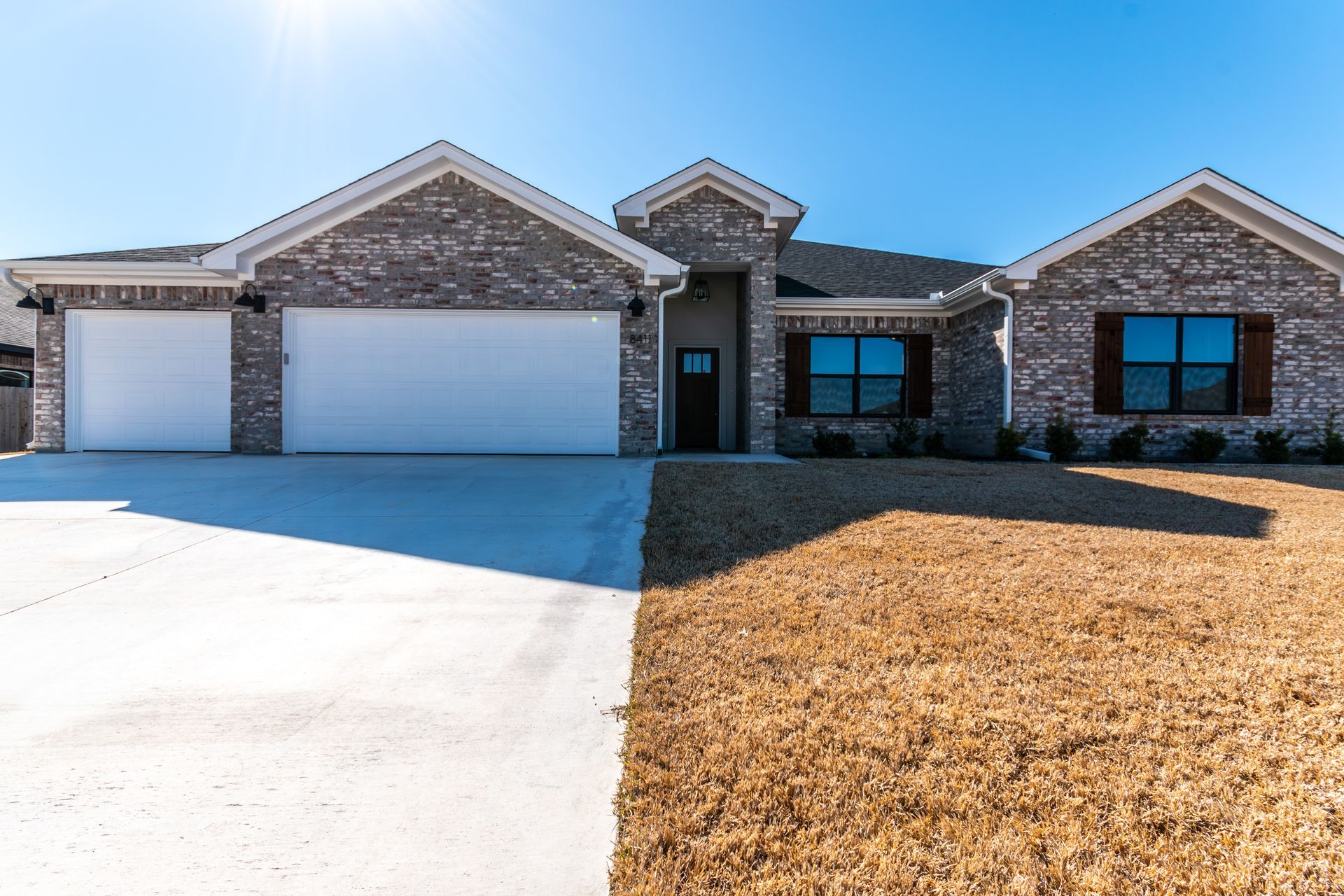 Brick house with two-car garage, front door, and dry brown grass lawn under a blue sky.