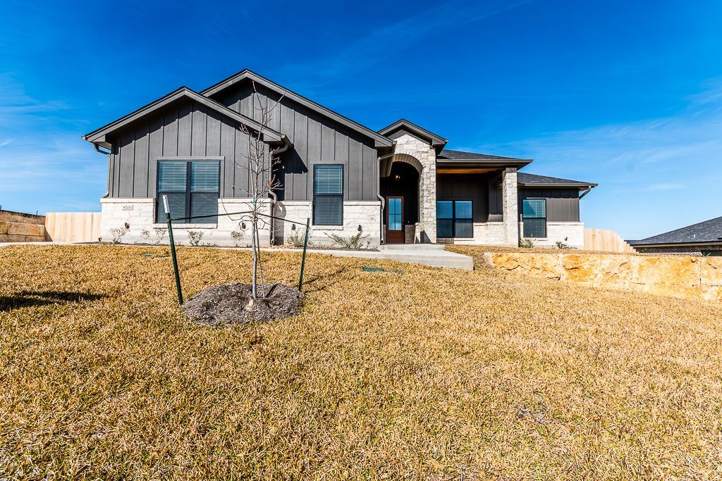 Modern single-story house with dark siding and stone accents under a clear blue sky, set behind a dry, grassy yard.