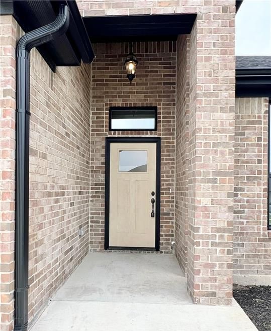 Tan front door with a window and black trim, brick facade, recessed entry, porch, and black rain gutter.