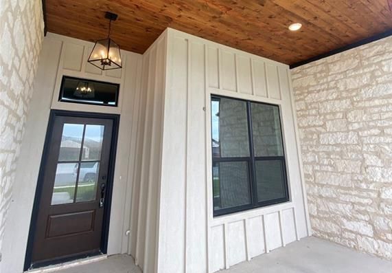 Covered porch with white vertical siding, stone wall, brown door and window, and wood ceiling.