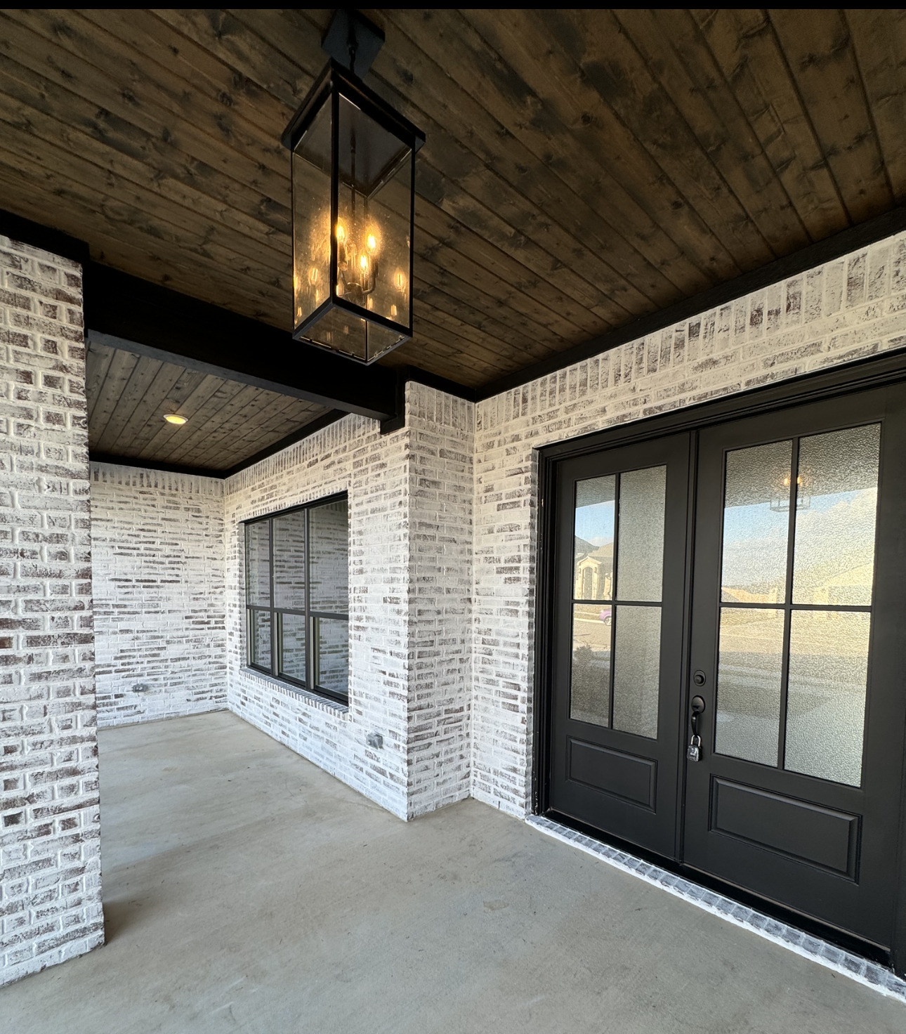 Covered porch with white brick walls, dark wood ceiling, black-framed windows and double doors, and a hanging lantern.