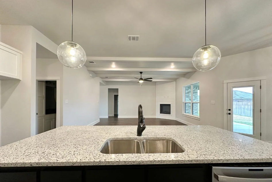 Kitchen with granite countertop island, sink, and pendant lights; view to living room with fireplace.