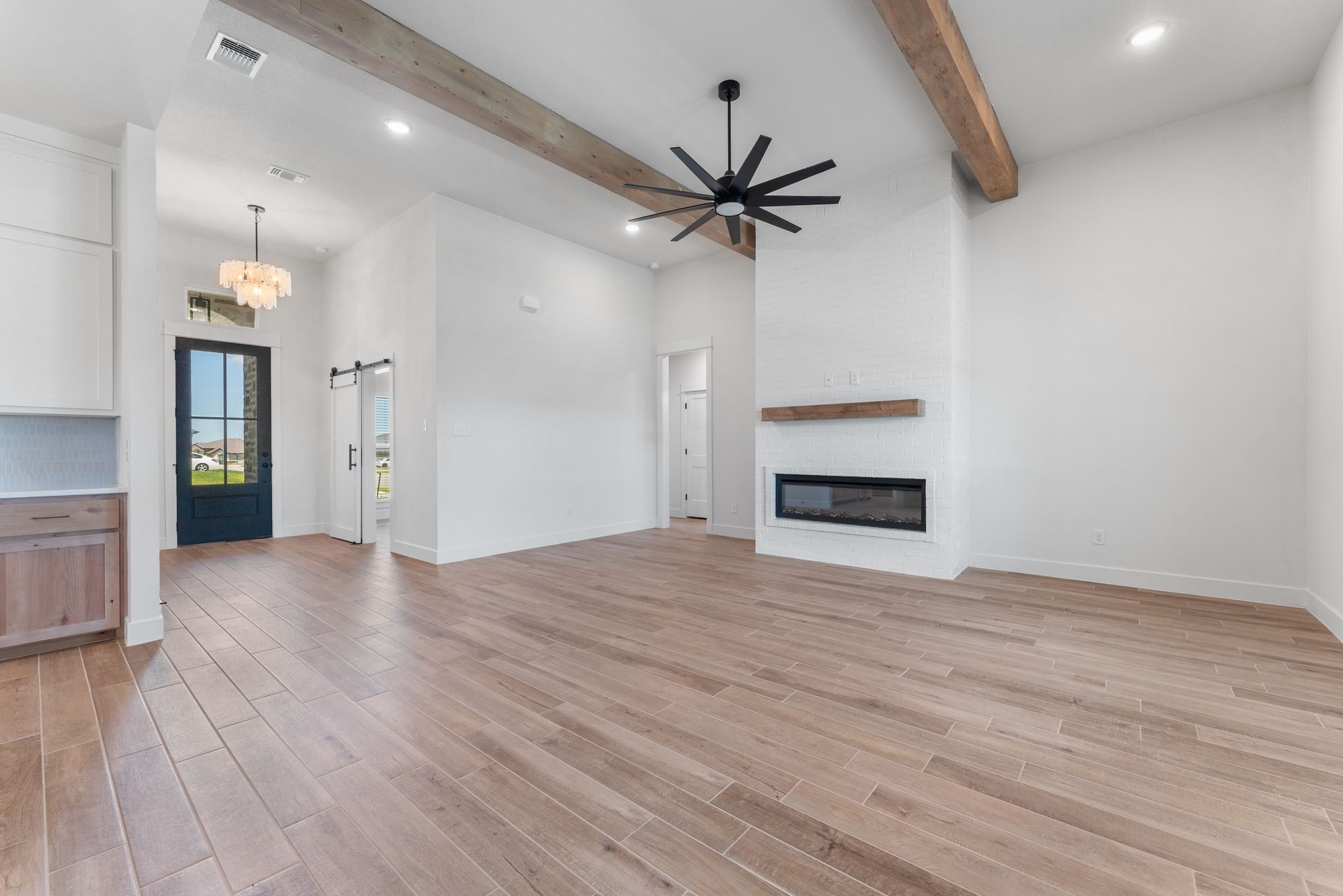 Spacious, empty living room with wood floors, white walls, and a fireplace. Exposed wood beams and a ceiling fan.