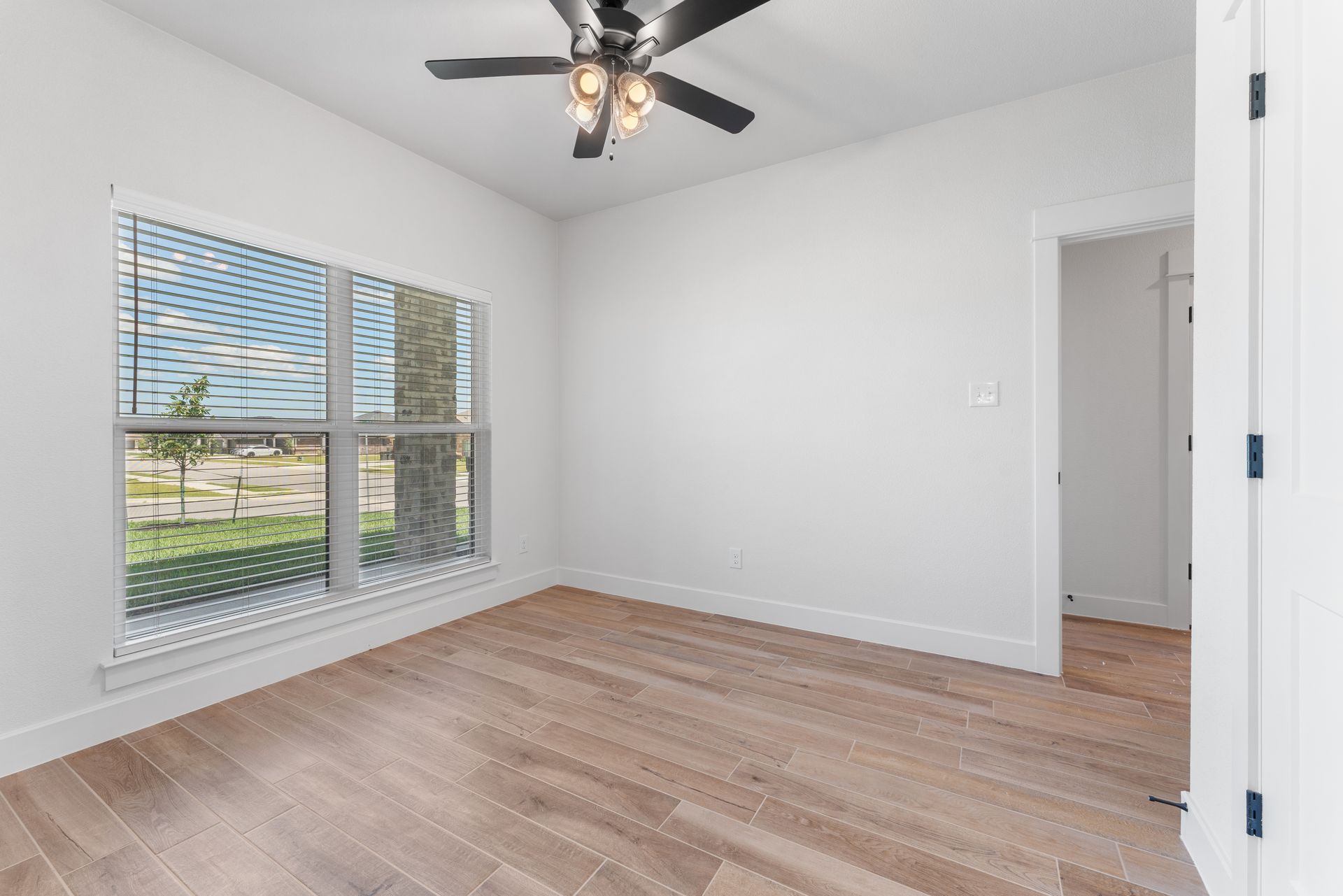 Empty room with wood floors, white walls, large window, ceiling fan, and open doorway.