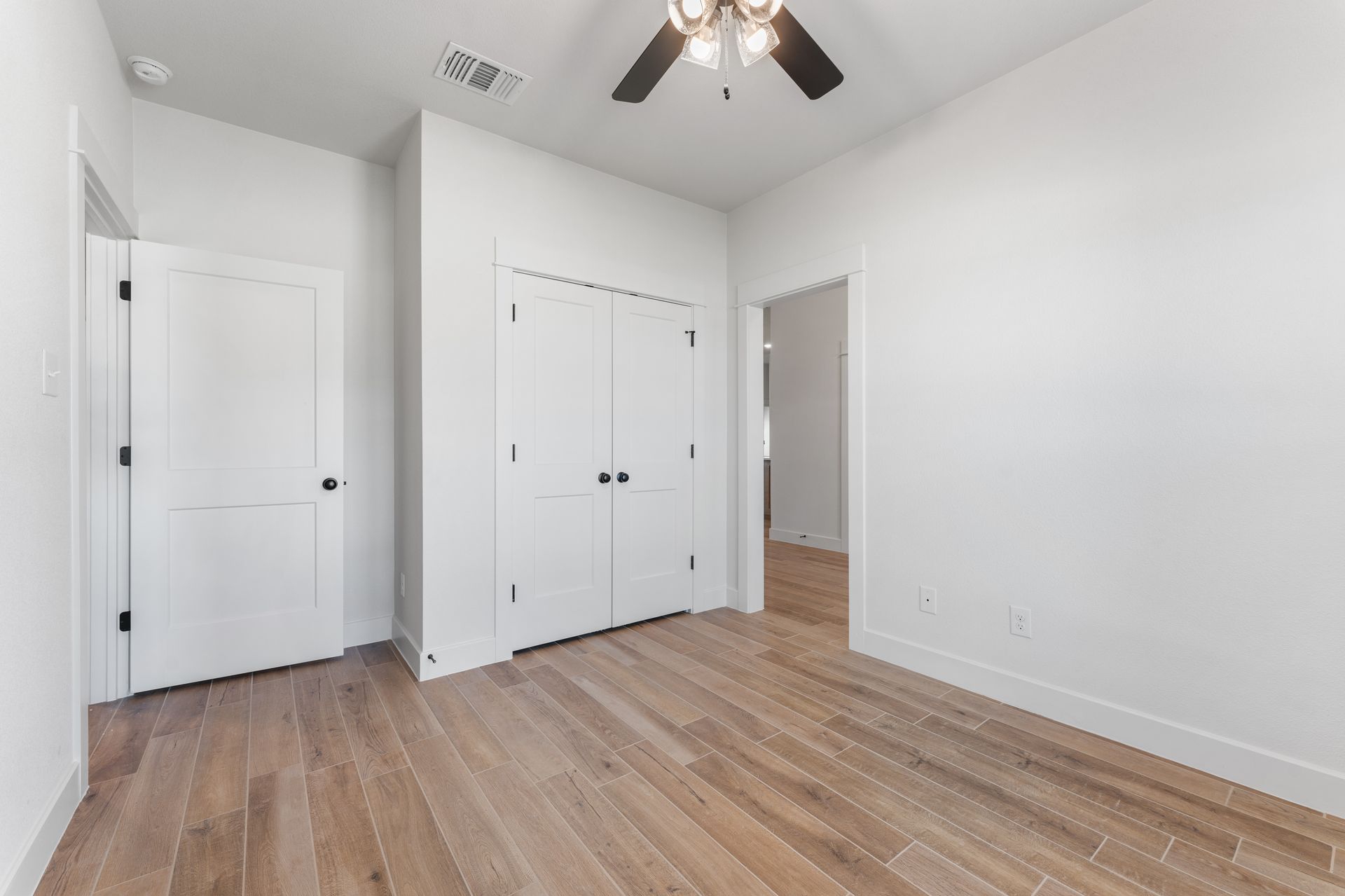Empty bedroom with wood floors, white walls, closet, and doors. Ceiling fan.