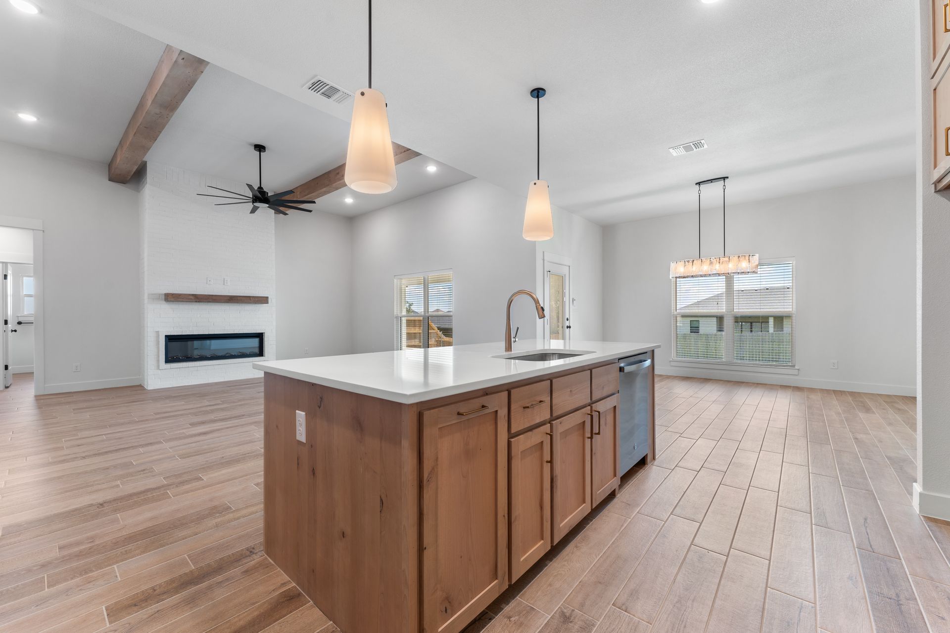 Spacious modern kitchen with island, wooden cabinets, and white countertops; wood floors, fireplace, and pendant lights.