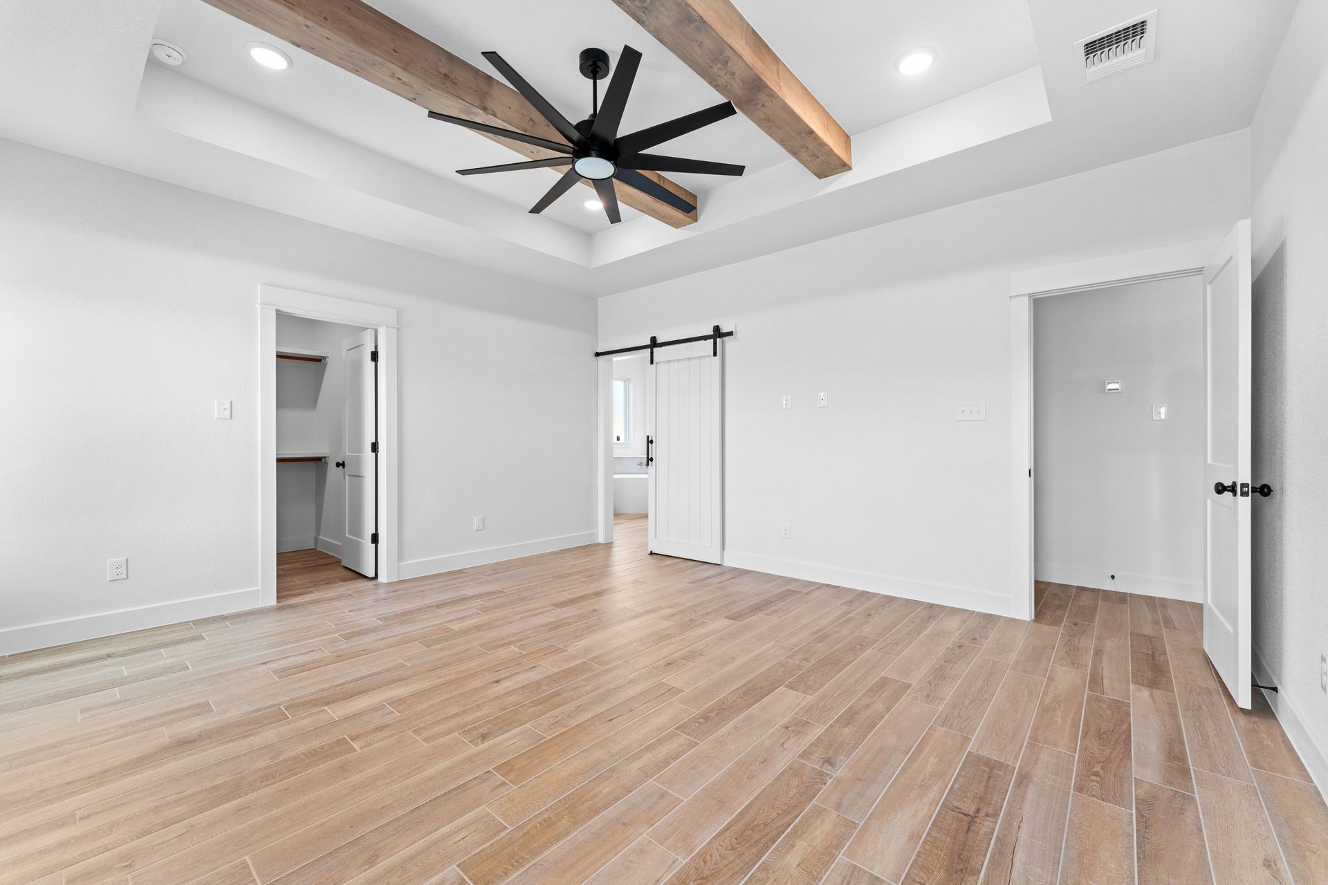 Spacious, white bedroom with wood floors, exposed beams, ceiling fan, and doors leading to a closet and bathroom.