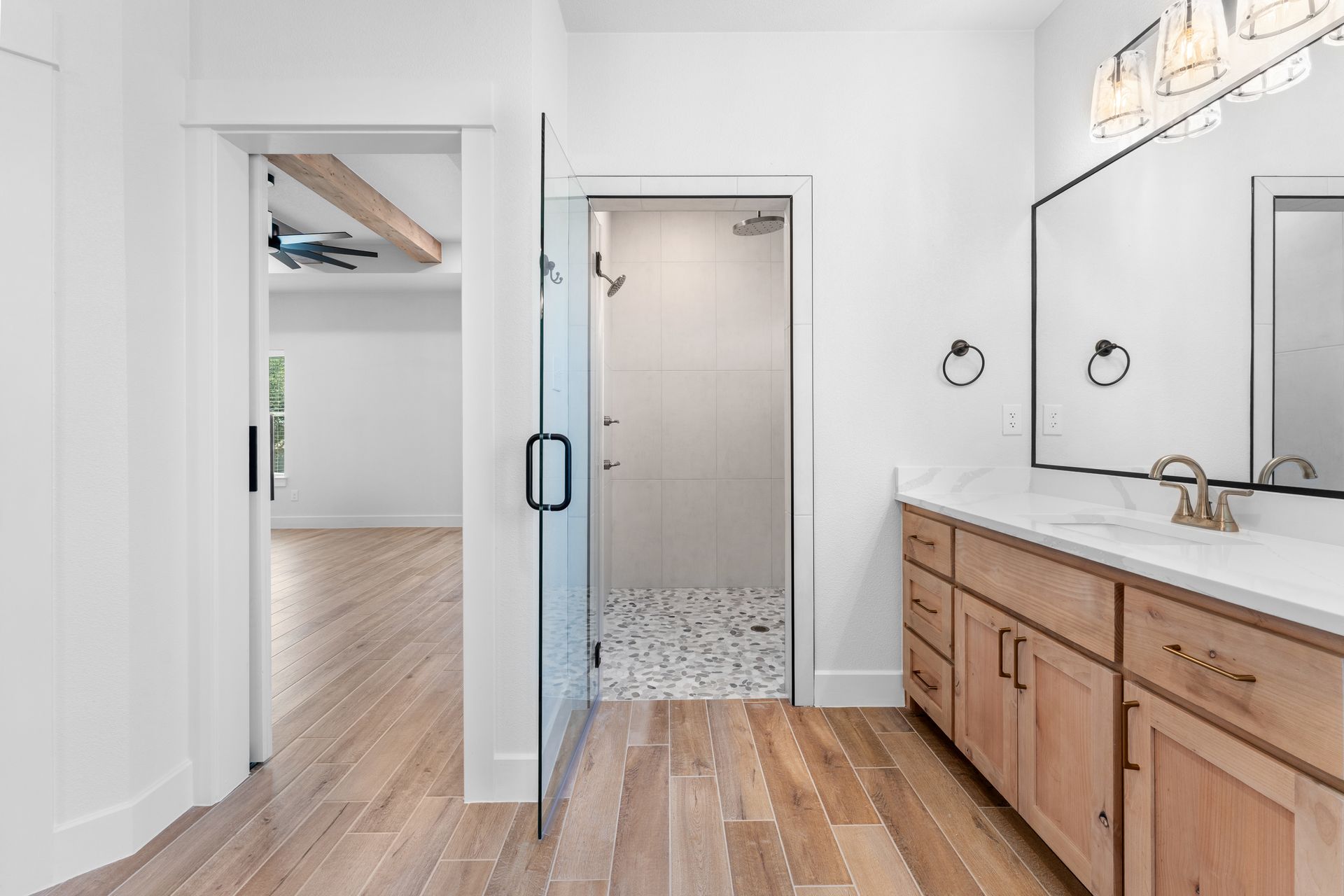Bathroom interior with a shower, vanity, and open doorway to a bedroom, with light wood floors and white walls.