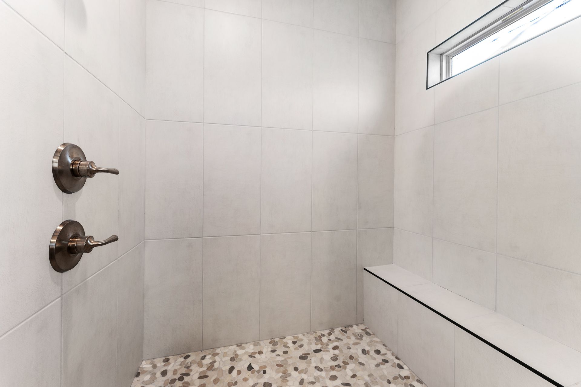 Empty shower with white tiled walls and a pebble floor, a built-in bench, and a small window.