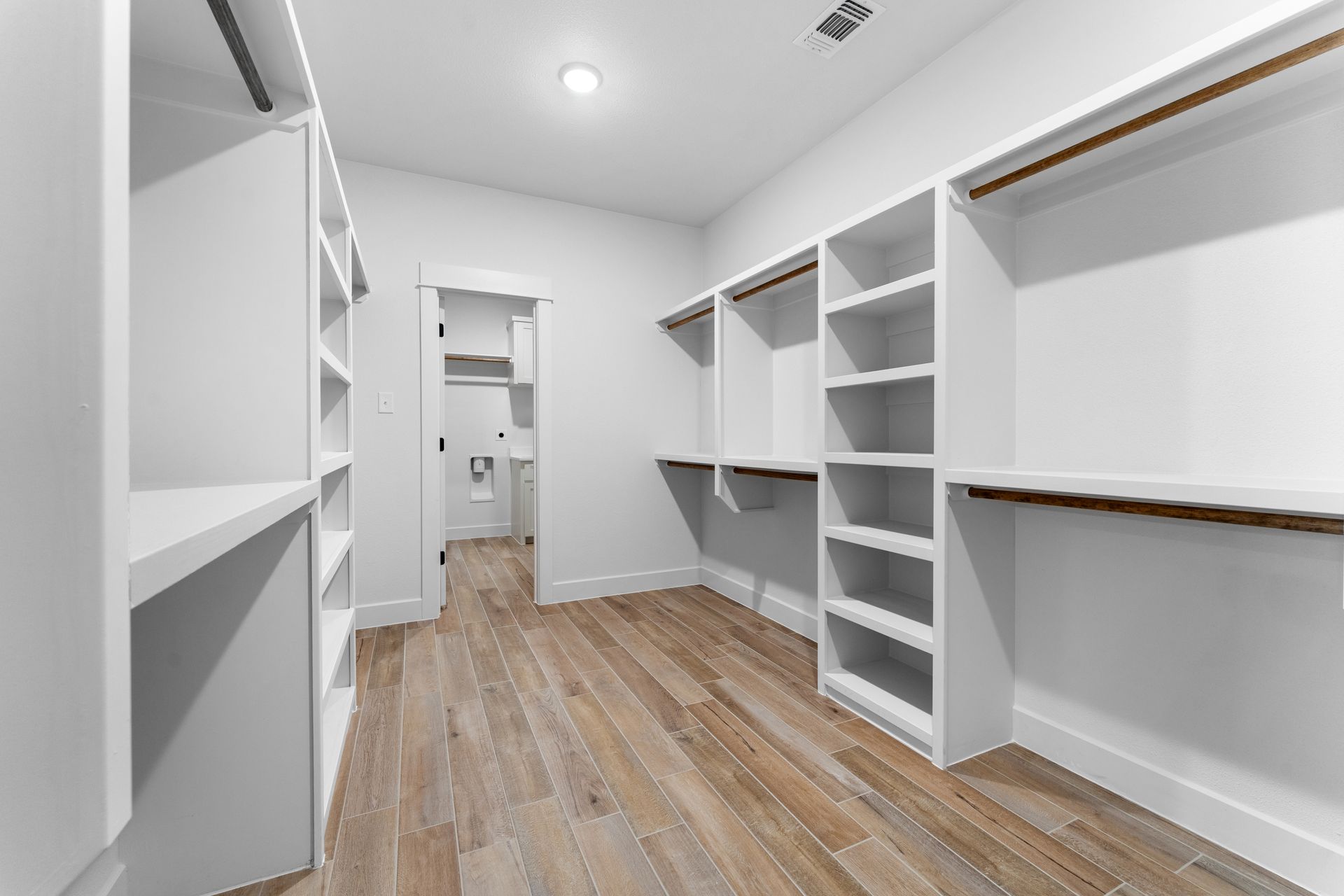 Empty walk-in closet with white shelves and wooden floor; a doorway leads to a bathroom.