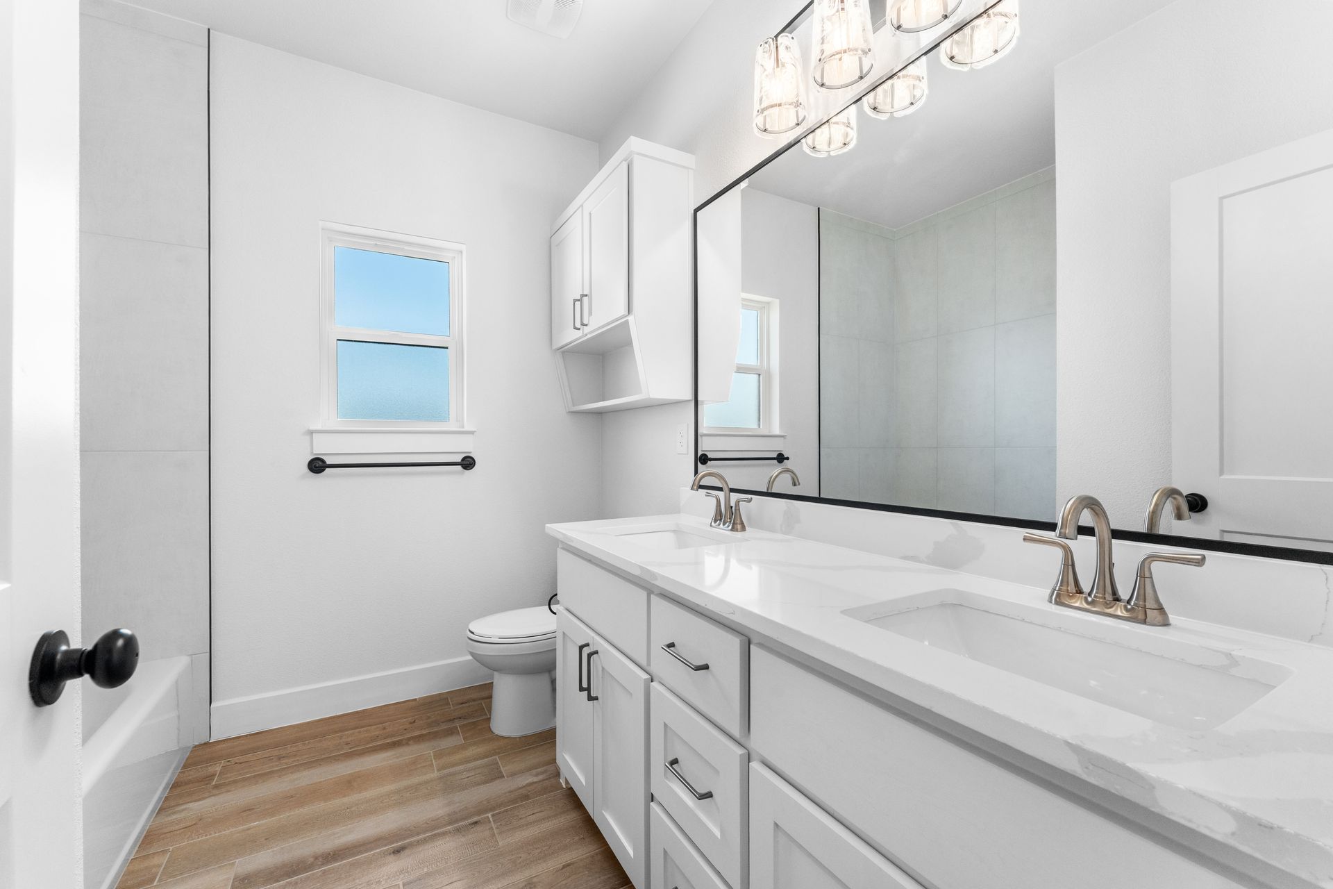 White bathroom with double vanity, large mirror, small window, and light wood floor.