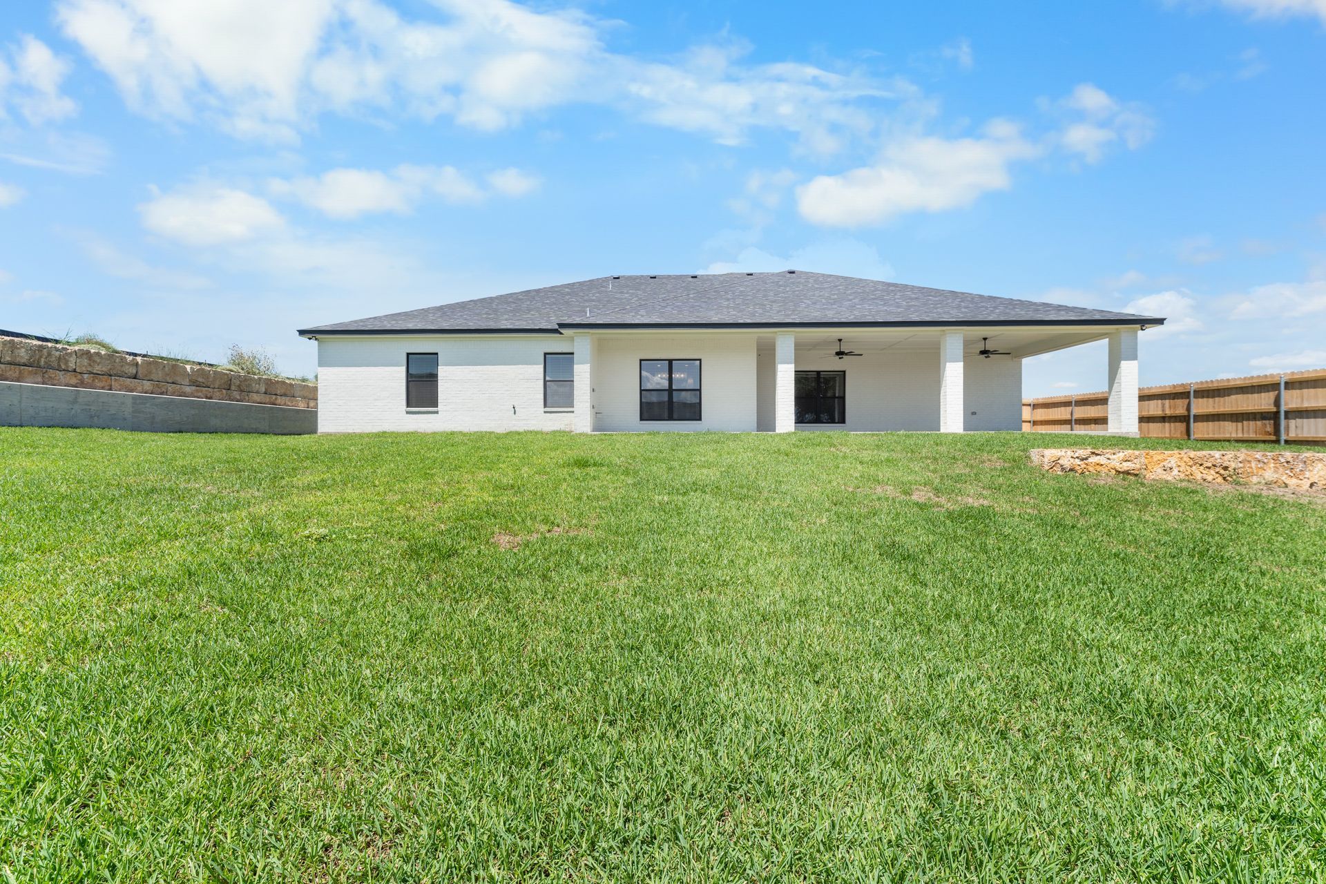 Backyard view of a single-story house with a covered patio, set on a grassy hill, under a blue sky.