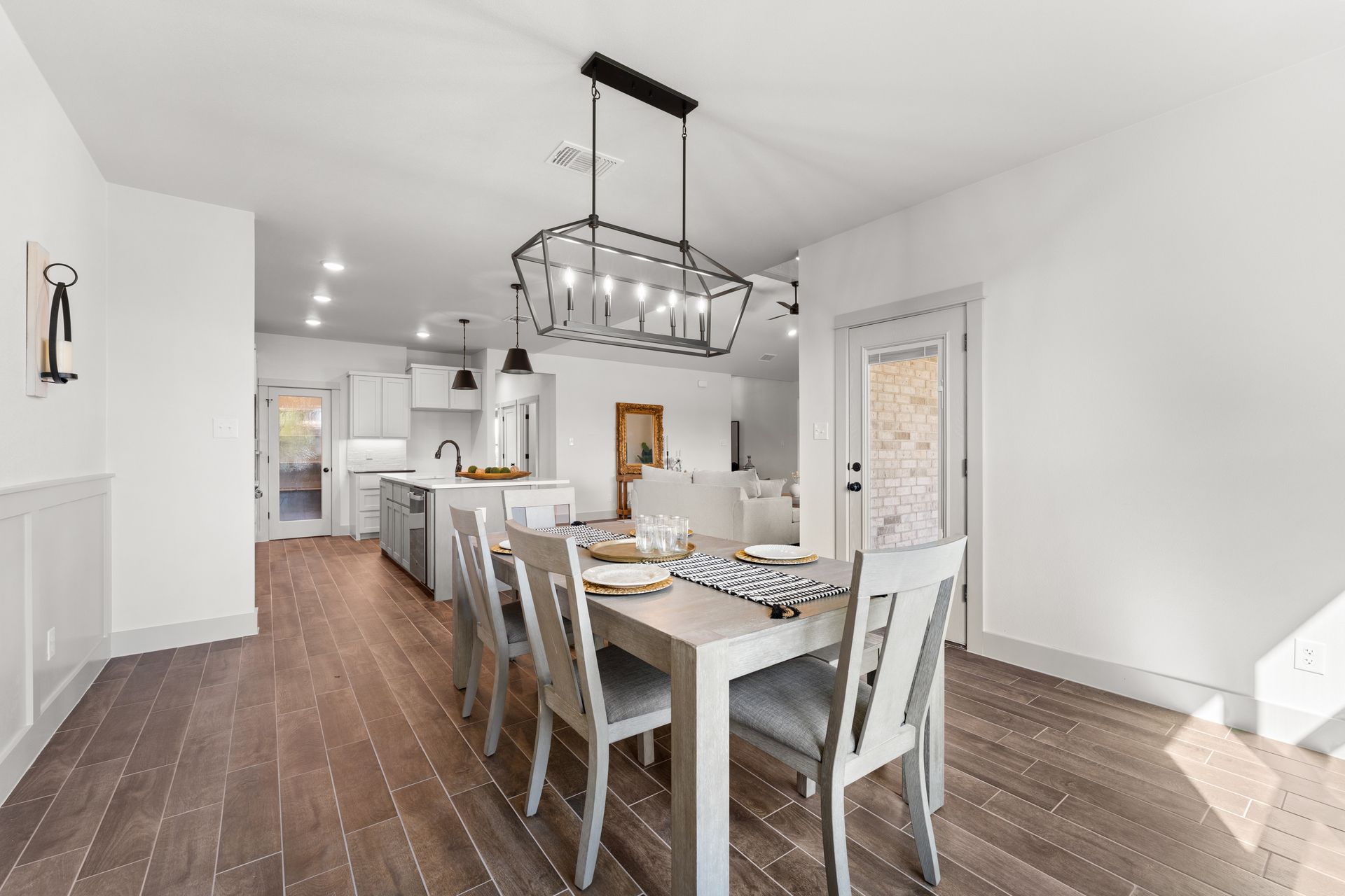 Dining room with table, chairs, and chandelier. Kitchen visible in the background. Wooden floors and white walls.