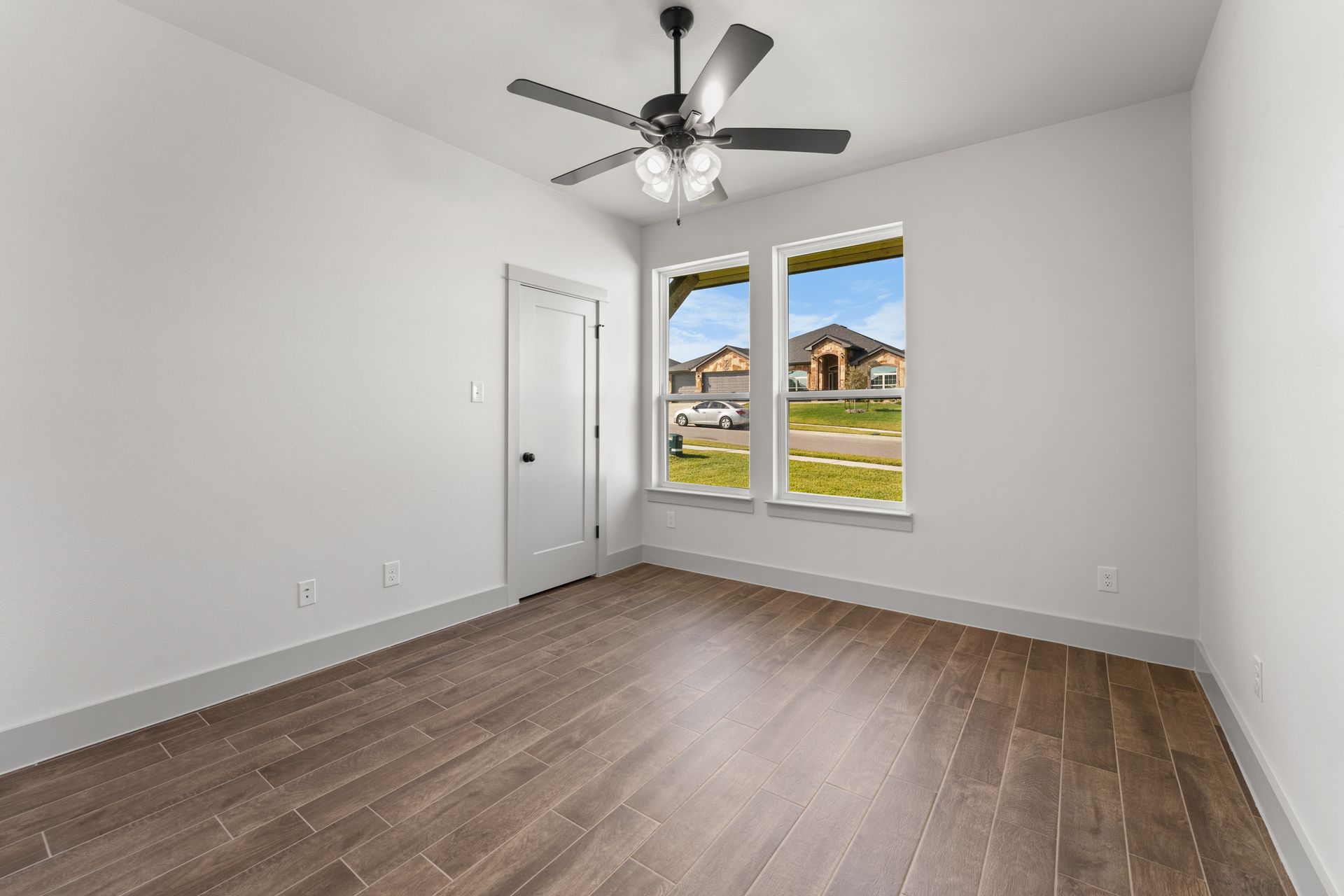 Empty bedroom with hardwood floors, white walls, and a ceiling fan; windows offer a view of a neighborhood.