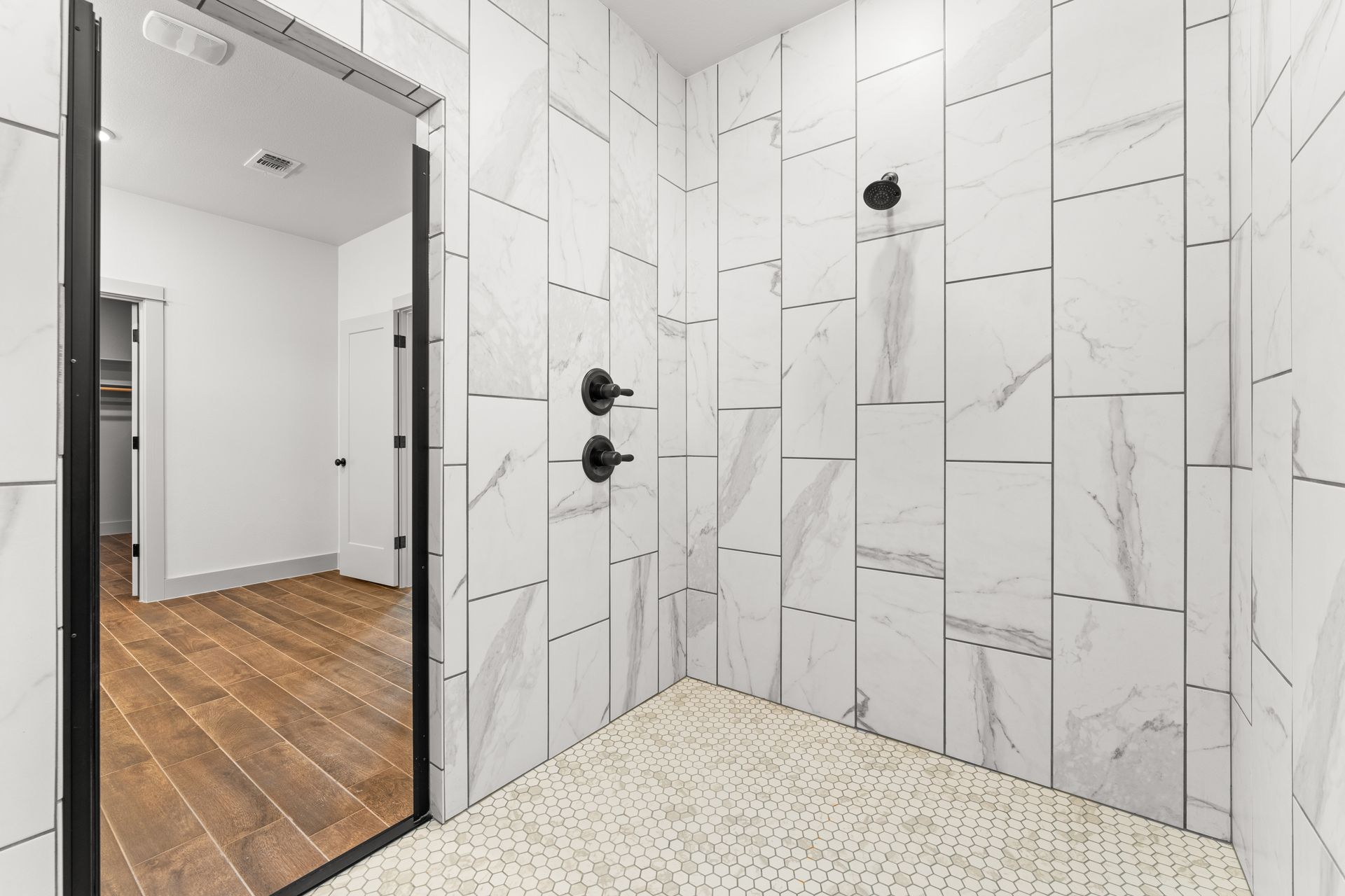 Modern bathroom with marble tiled shower, black fixtures, and wooden floor visible through open door.