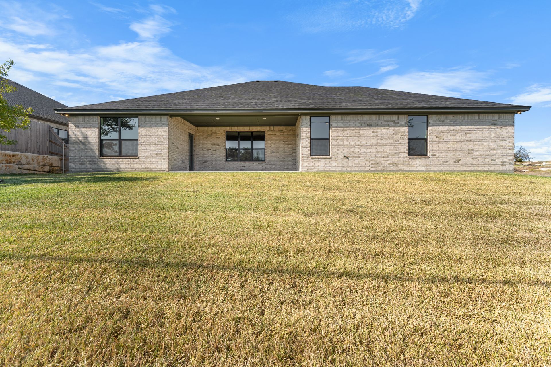 Back of a one-story brick house with a dark roof and windows, set in a grassy yard under a blue sky.