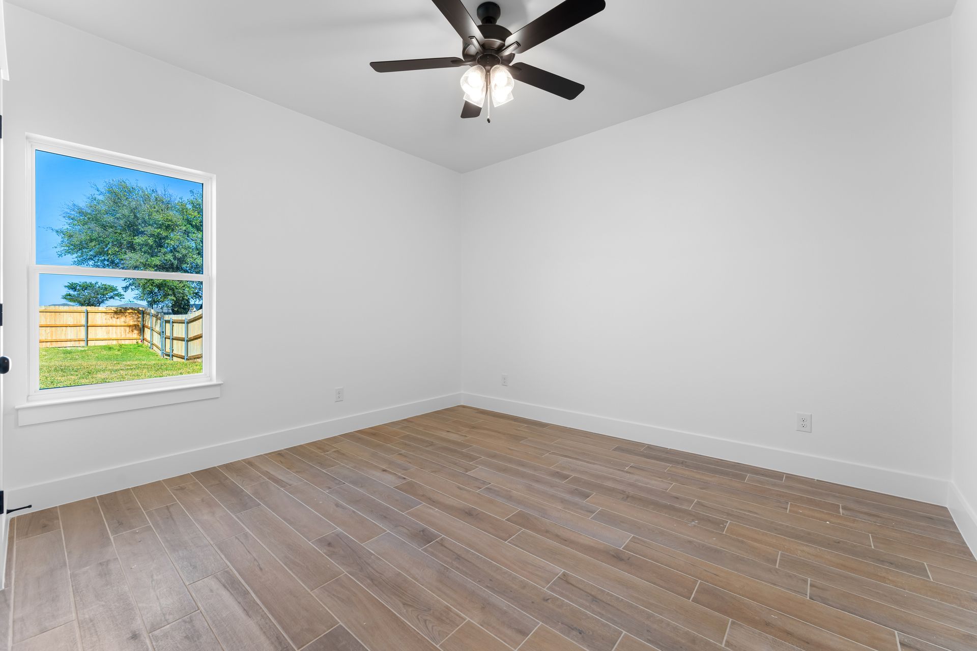 Empty room with wood-look floor, white walls, ceiling fan, and window overlooking a yard with a fence.