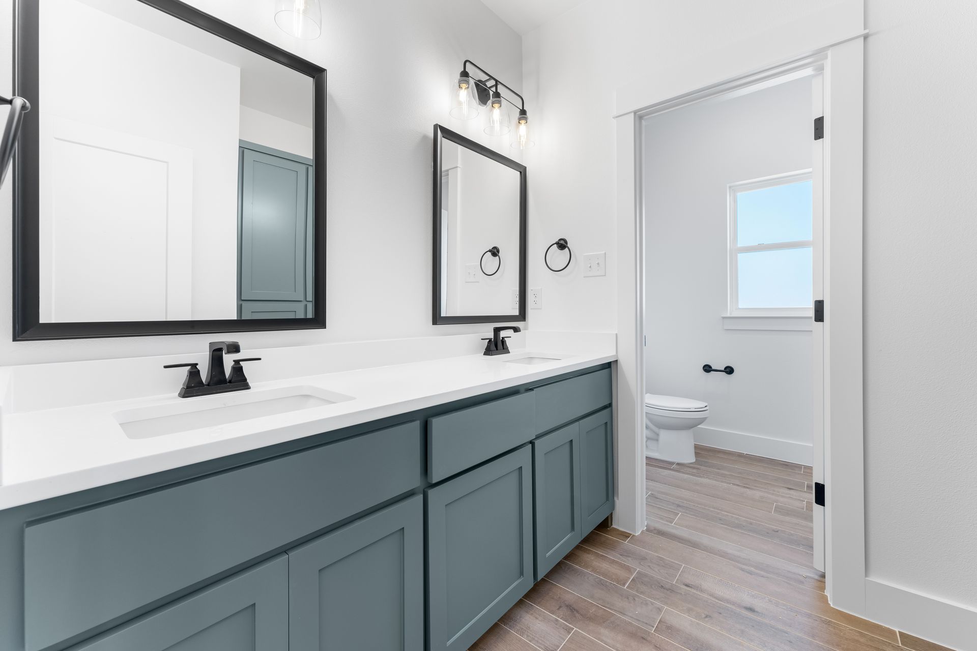 Bathroom with dual sinks, teal cabinets, black framed mirrors, and a doorway to a toilet.