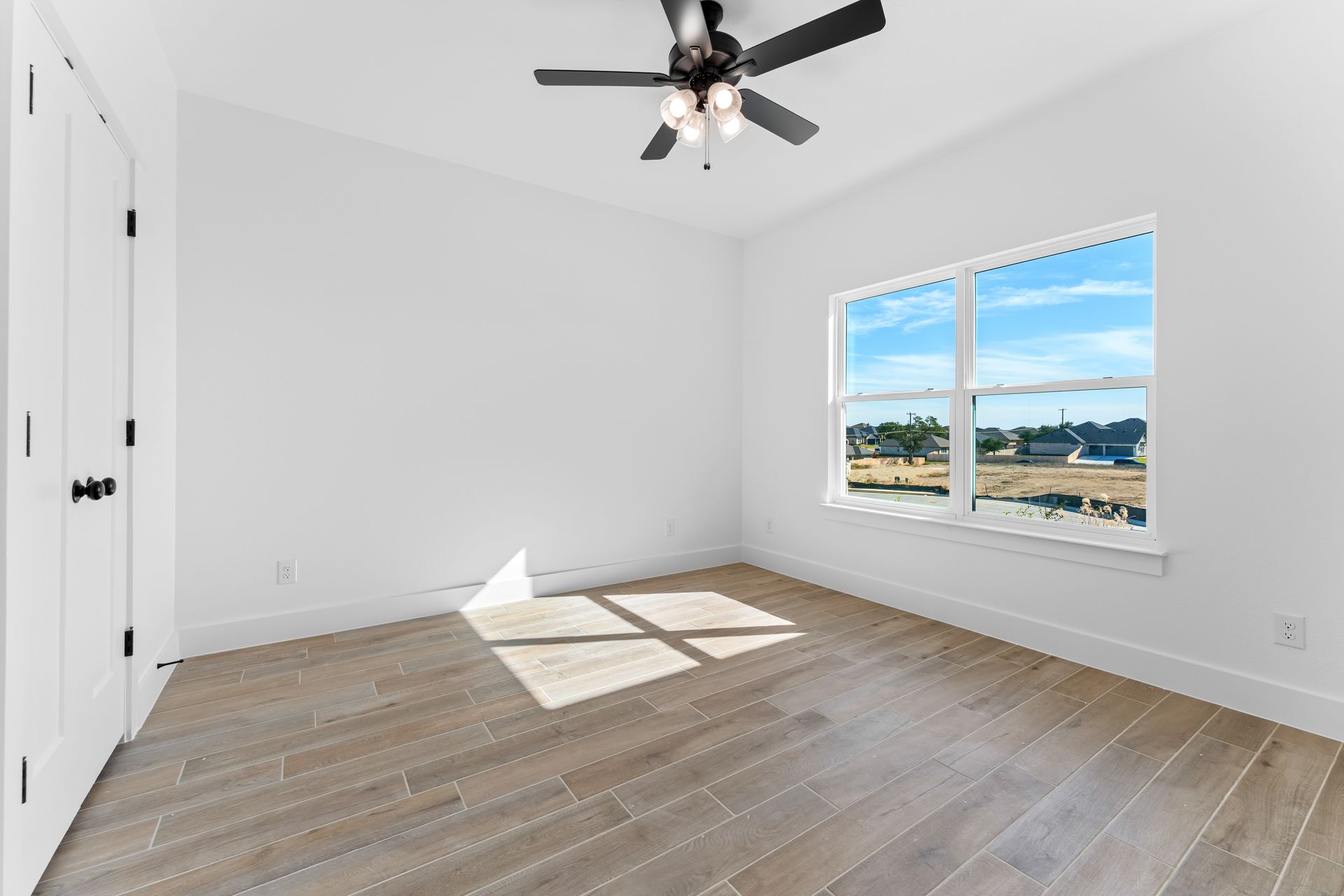 Empty room with light wood floor, white walls, window with view, and ceiling fan.