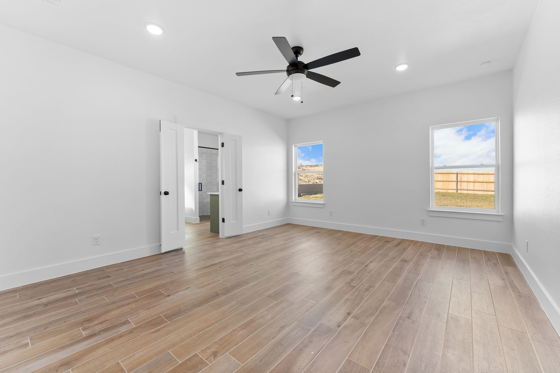 Empty bedroom with light wood floors, white walls, two windows, and a ceiling fan.