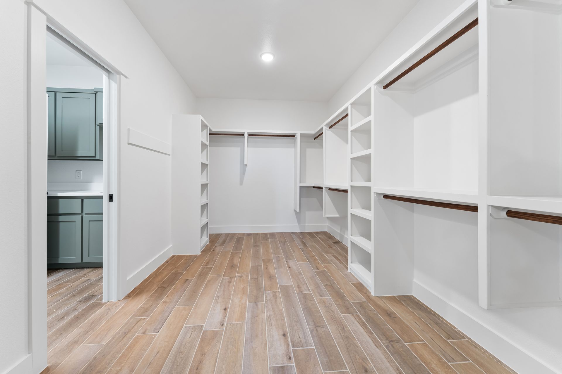 Walk-in closet with white shelves and wooden floor. A doorway reveals a kitchen.