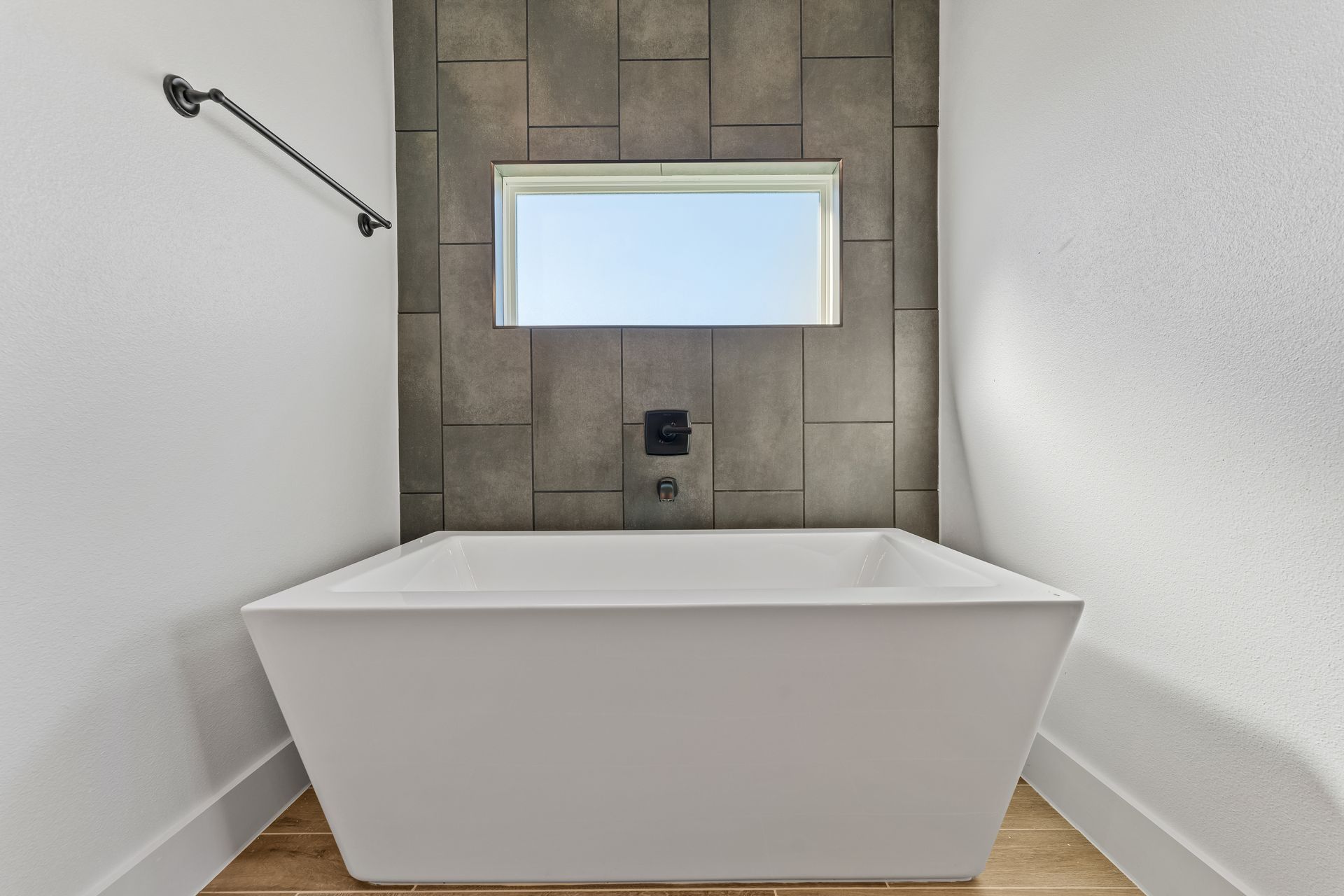 A white, modern bathtub in a bathroom with a window and gray tiled accent wall.
