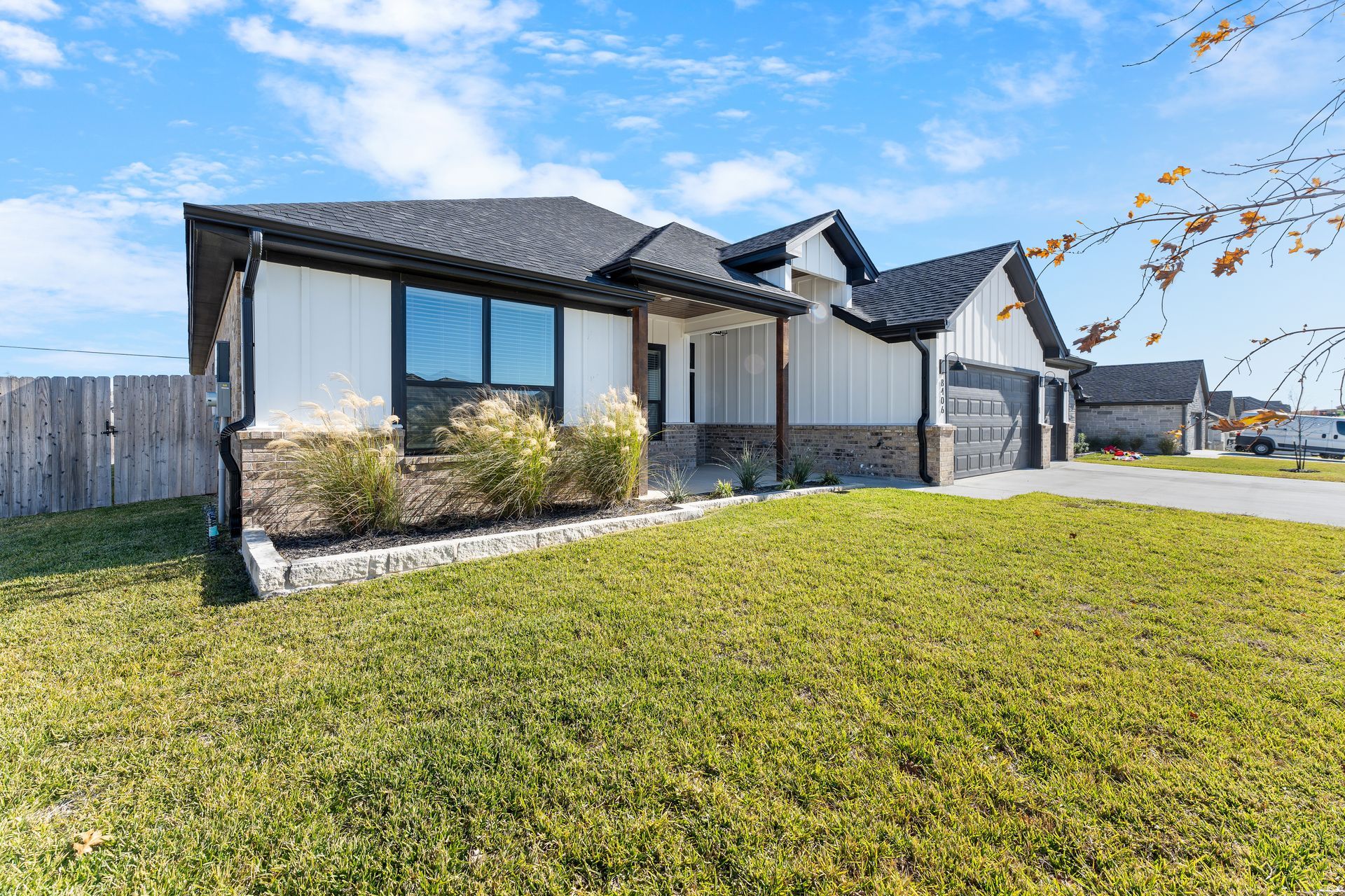 A white house with a black roof, brick accents, and a garage, set on a grassy lawn under a blue sky.