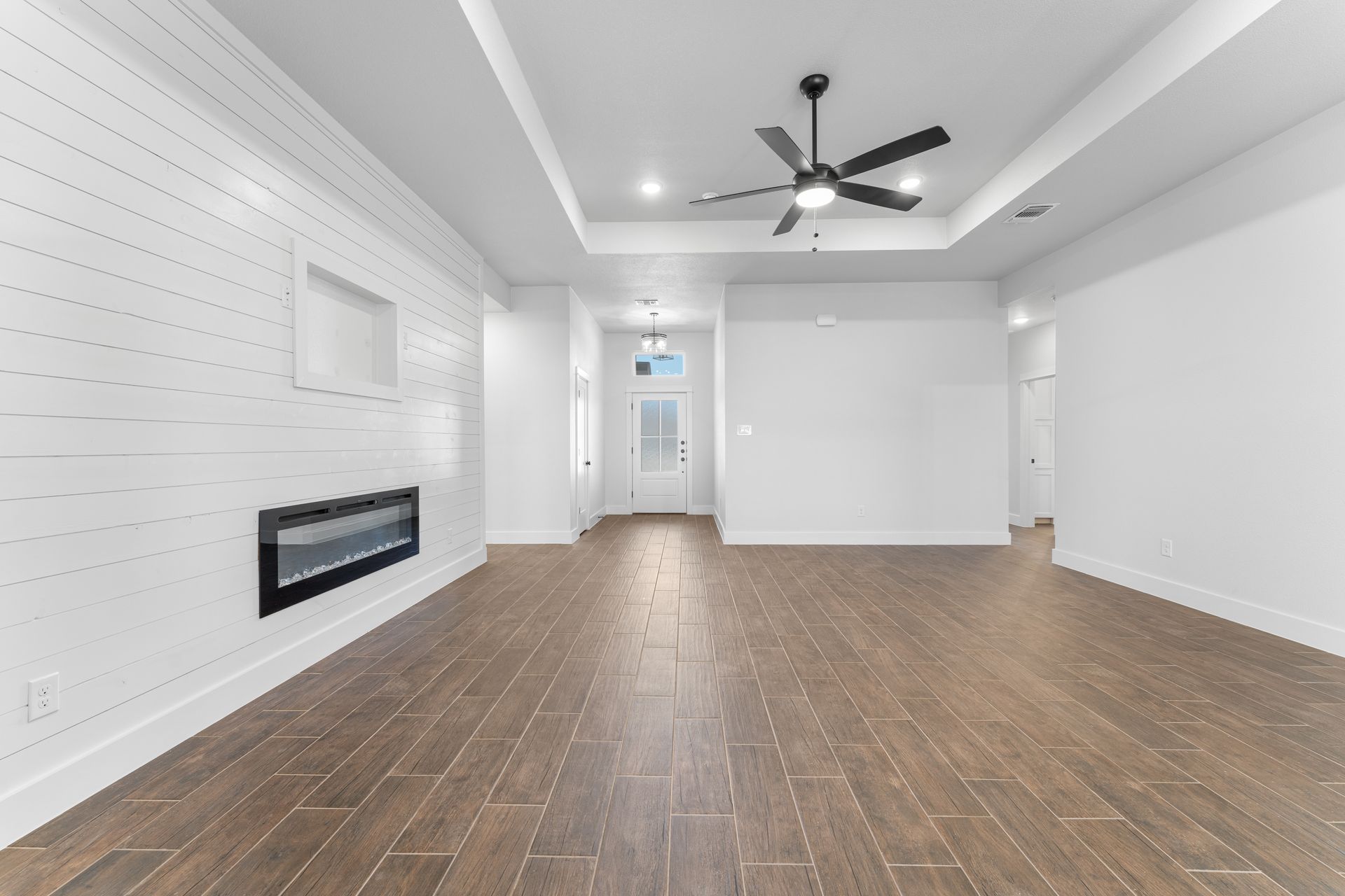 Empty living room with wood flooring, white walls, recessed ceiling, fireplace, and ceiling fan.