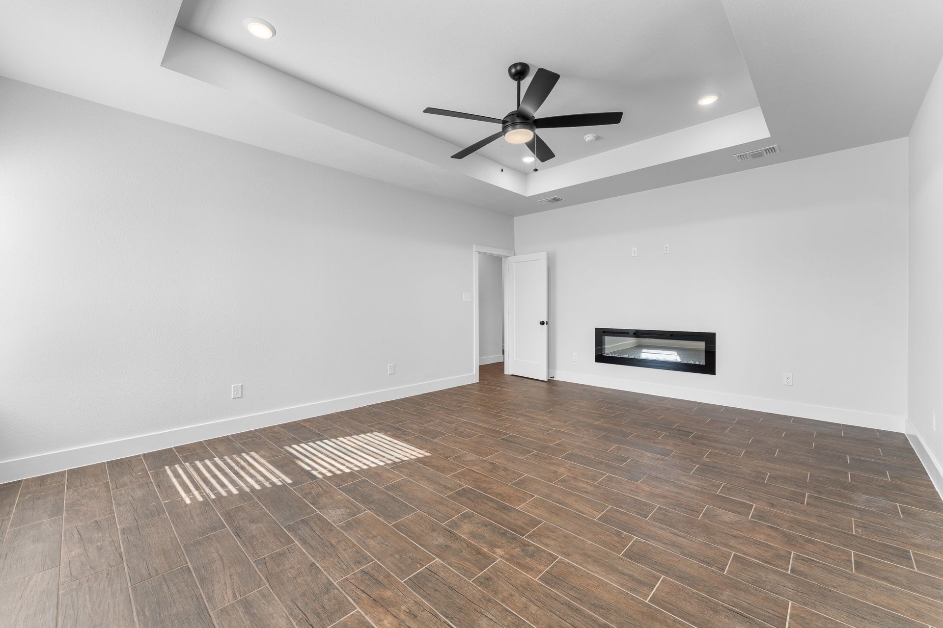 Empty room with wood floors, recessed ceiling with fan, fireplace, and white walls.