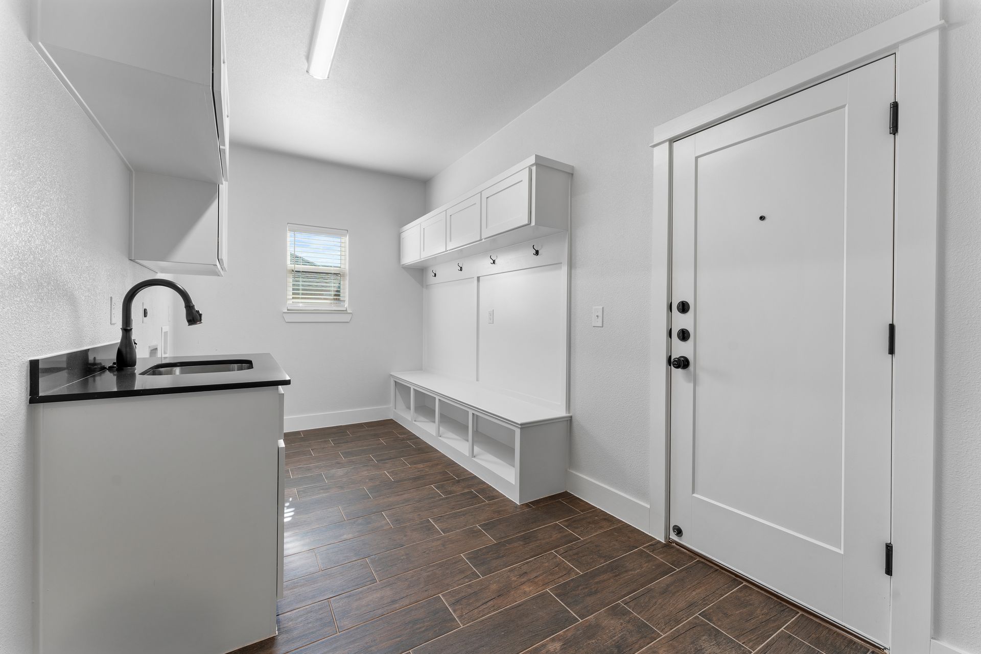 Laundry room with white cabinets, dark sink, coat rack, bench, and door. Brown tile floor.