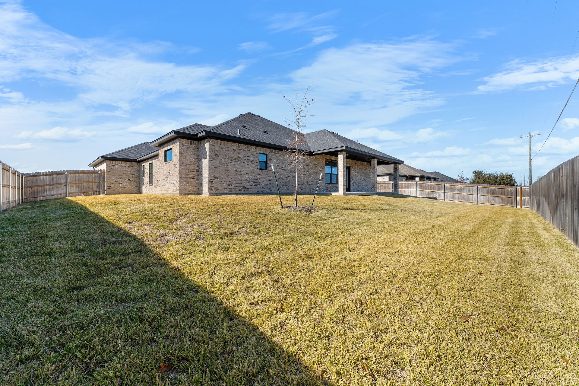 A brick home with a sloping grassy yard, under a blue sky. A wooden fence surrounds the property.