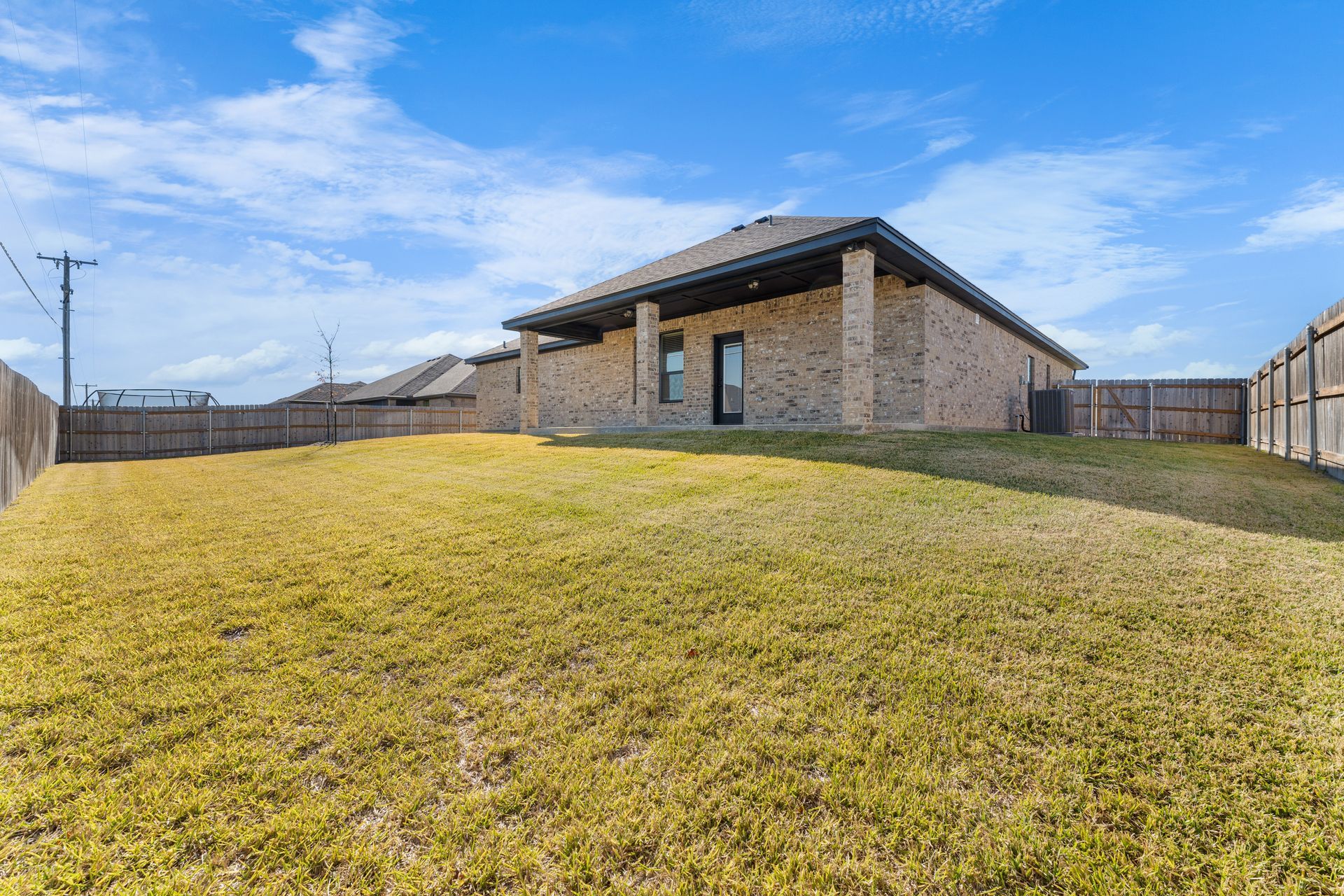 Backyard of a brick house with a brown wooden fence, green grass, and a blue sky.