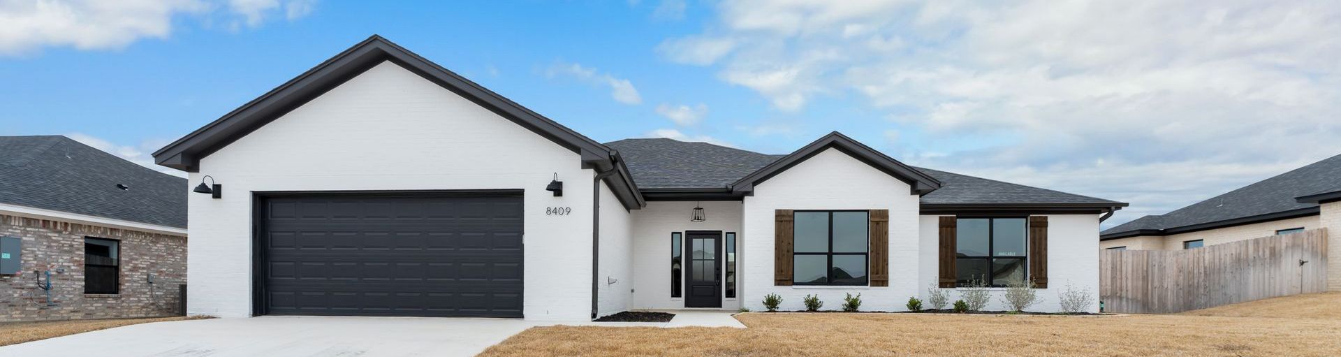 White house with black roof and garage door, surrounded by dry grass and other houses. Blue sky.