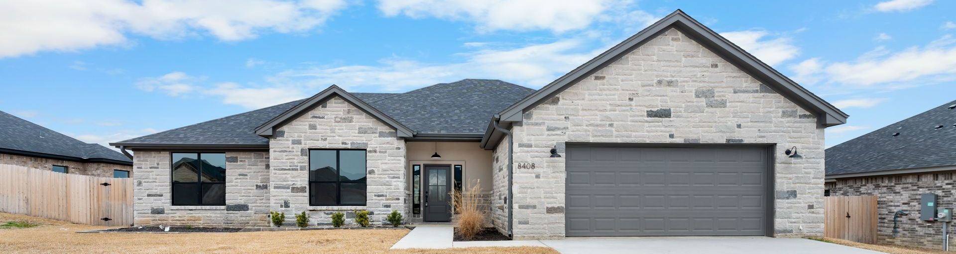 A new stone home with a gray roof and a two-car garage under a blue sky with clouds.
