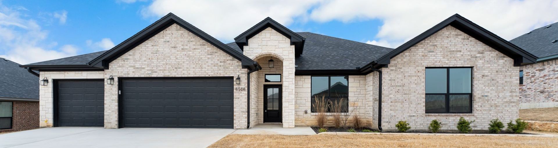 A beige brick house with a dark roof and two-car garage under a cloudy sky.
