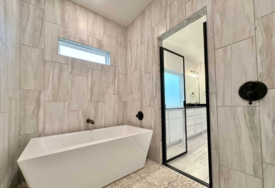 Modern bathroom with white tub, stone-like tiled walls, and black-framed glass door leading to a vanity.