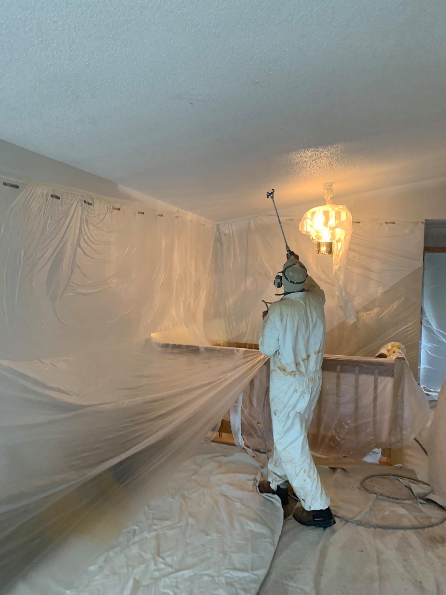 Person in white coveralls sprays paint on a room wrapped in plastic sheeting.