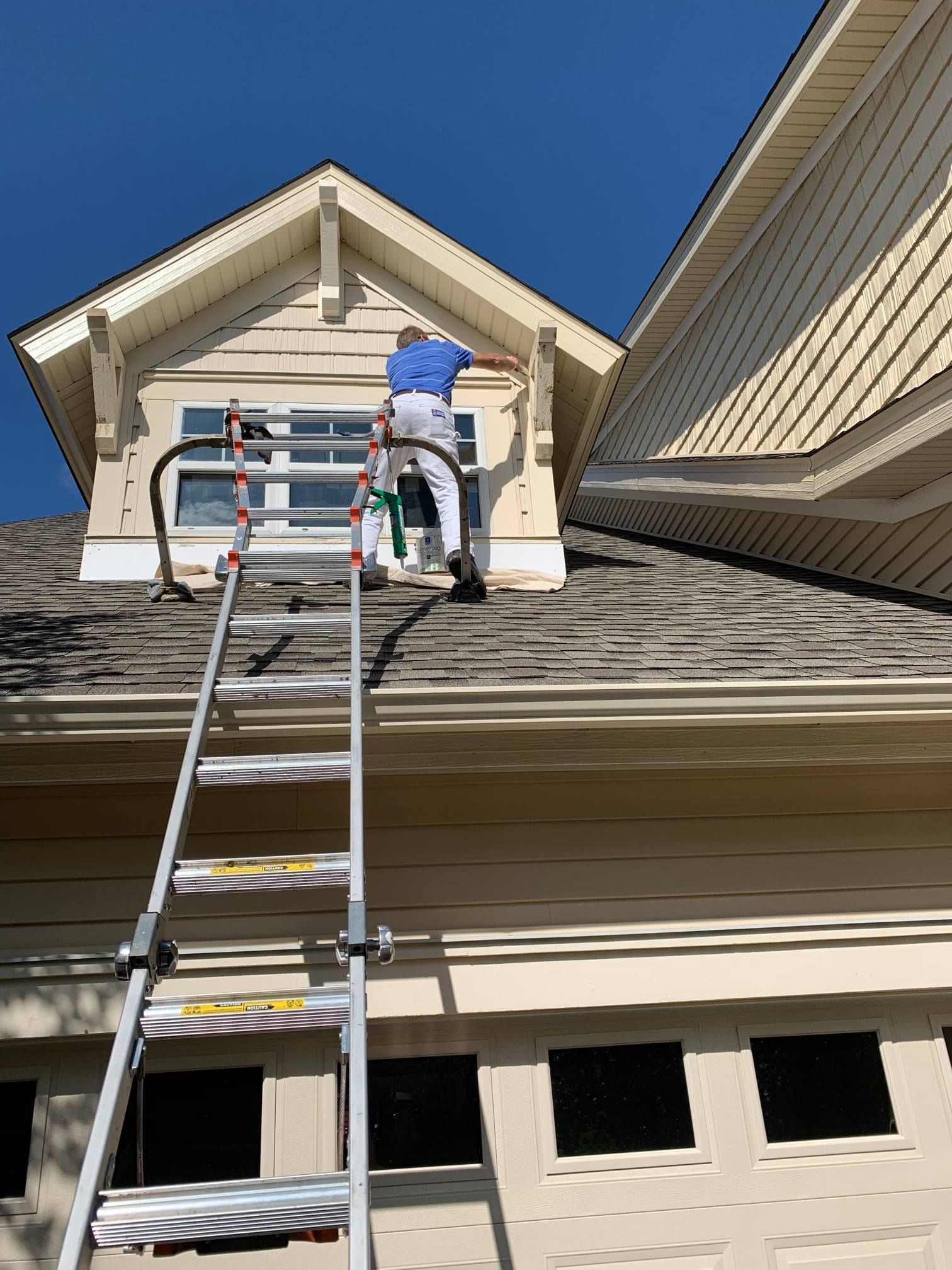 Person on a ladder repairing a white house gable under a clear blue sky