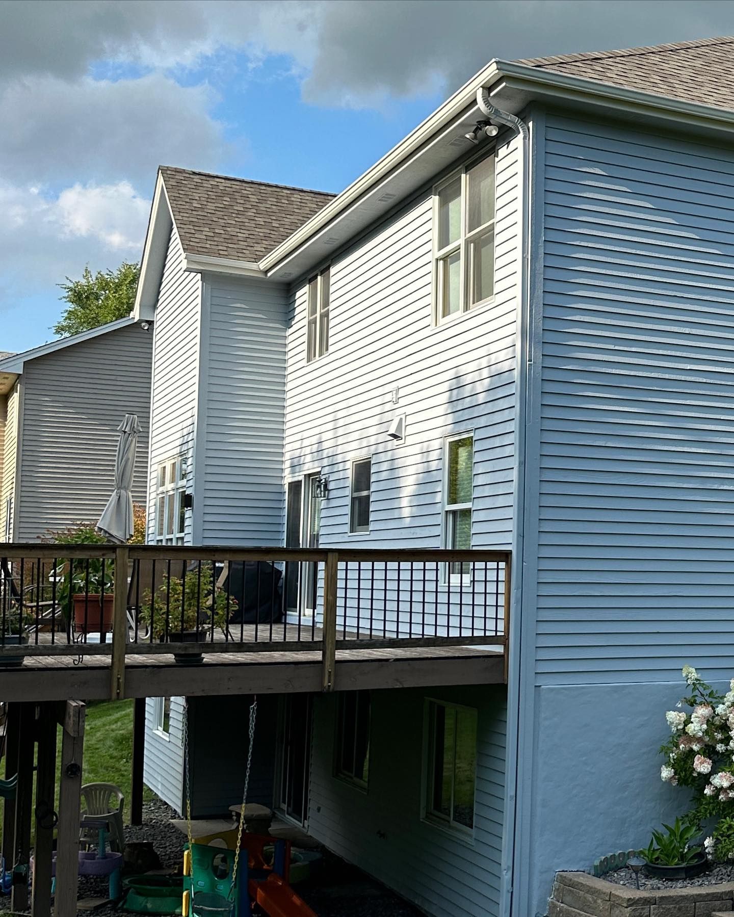 Two-story light blue house with a wooden deck and stairs, seen from the side under a cloudy sky