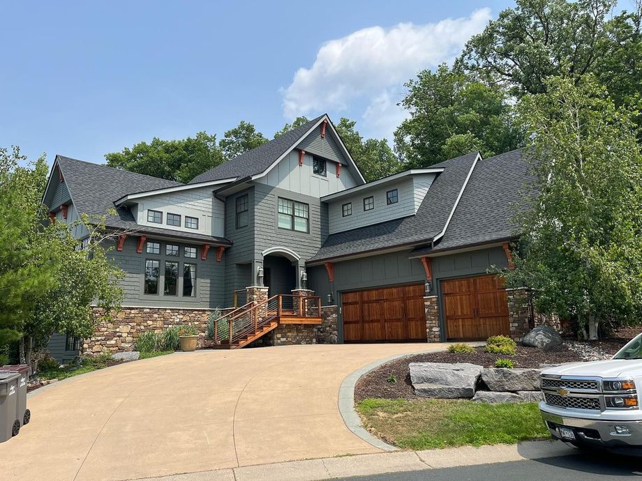 Large gray stone house with wood garage doors, curved driveway, and trees under a blue sky
