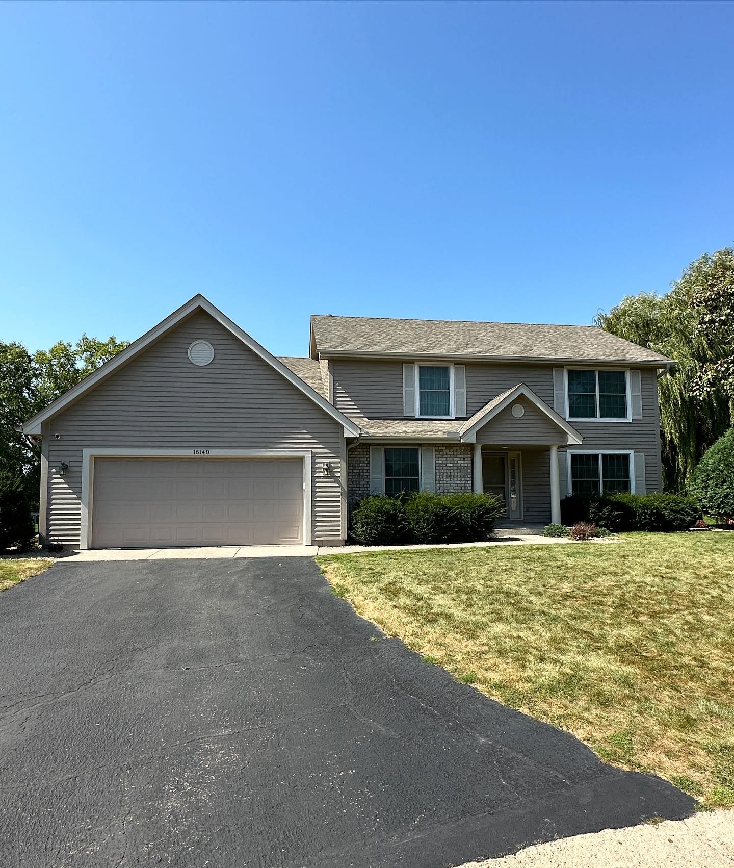 Two-story suburban house with gray siding, white garage, black driveway, and a sunlit lawn under a clear blue sky