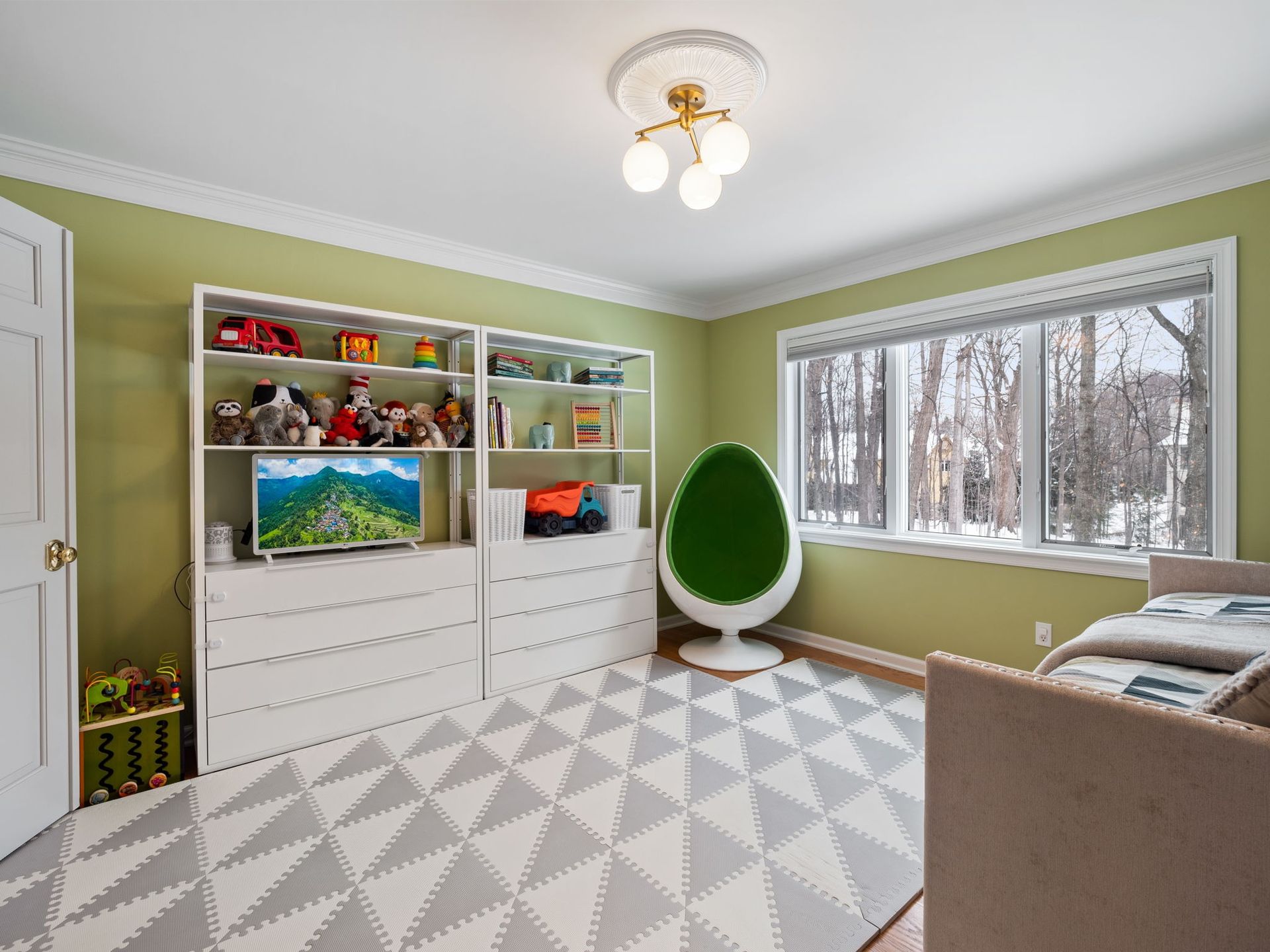 Bright playroom with white shelves, geometric rug, green chair, and large window with snowy trees outside