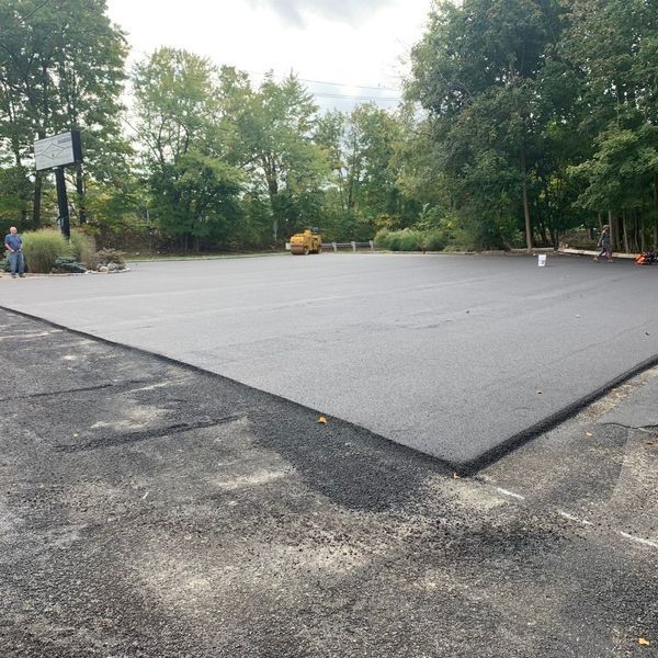 Freshly paved asphalt parking lot; trees in the background, a person standing at the edge.