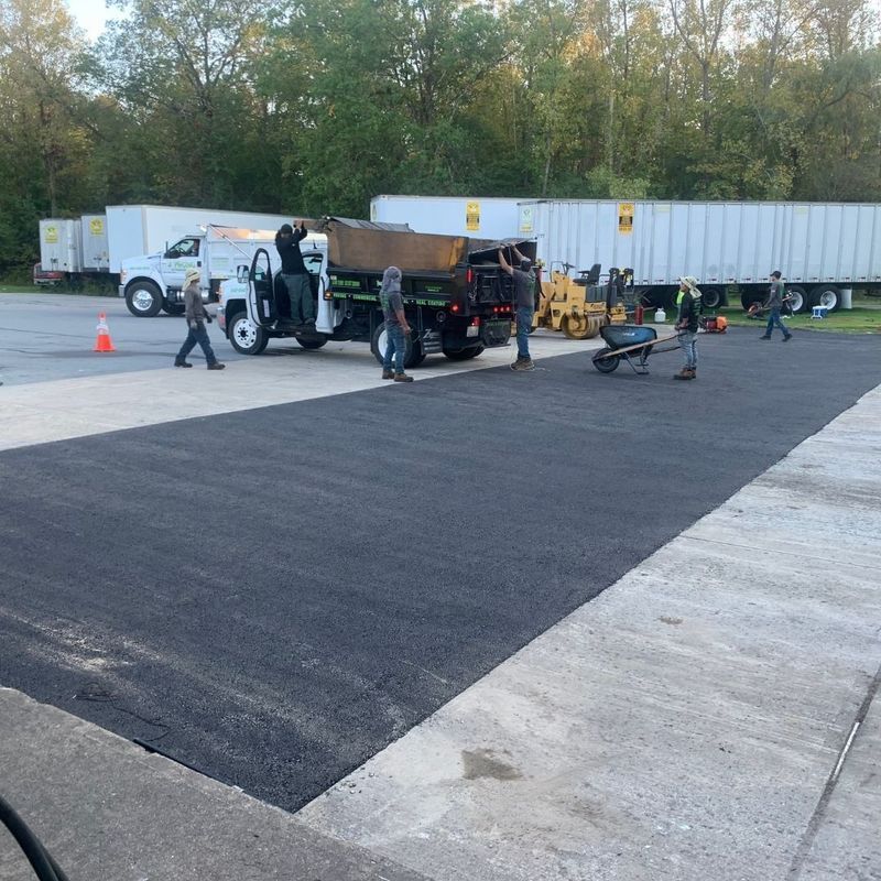 Workers paving asphalt in a parking lot, with a truck, wheelbarrows, and white trailers in the background.