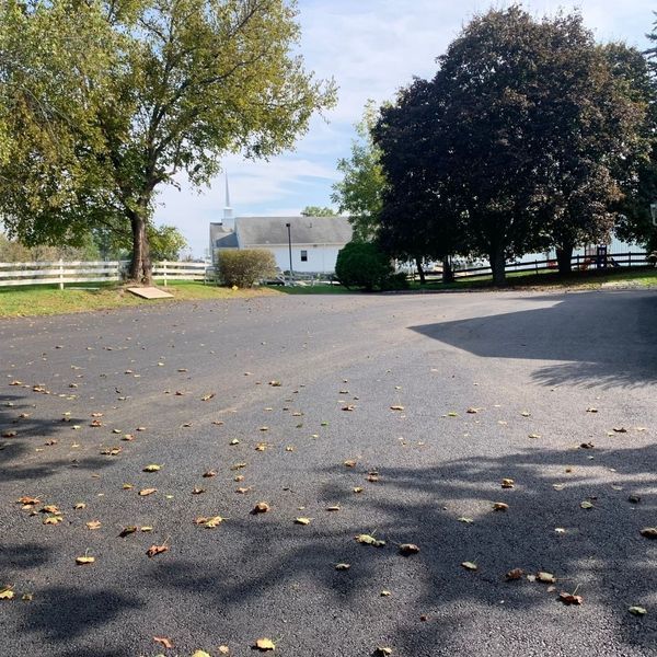Asphalt parking area with scattered leaves, leading to a white building with a steeple, trees and a fence.