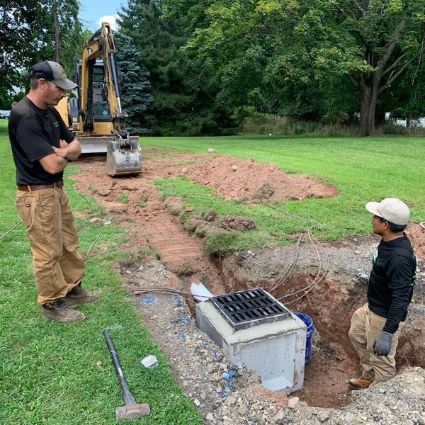 Two workers by an excavated drainage unit with an excavator in the background.